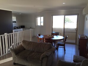 A living room with a sofa, a dining table with chairs, a kitchen area with cabinets and a stove, and a balcony with a railing. - Inverloch Cabins (Inverloch)