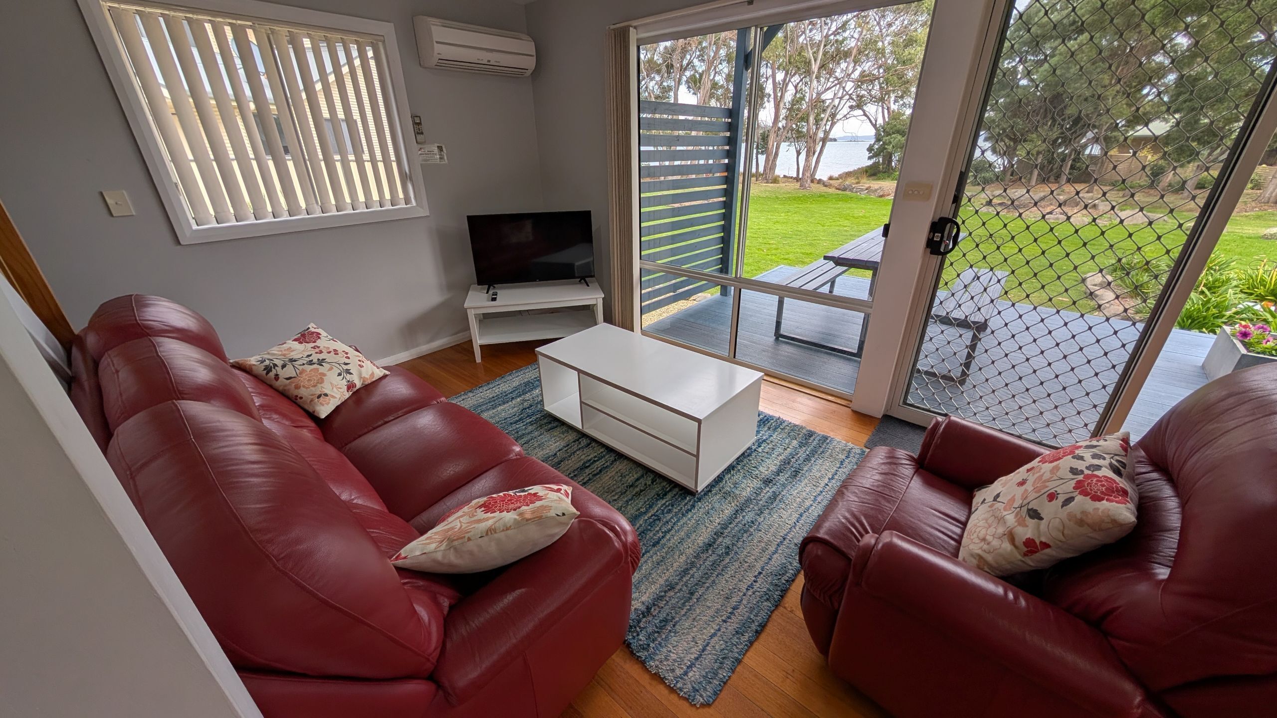 A living room with two red leather sofas, a white coffee table, a flat-screen TV on a stand, and a sliding glass door leading to an outdoor patio.