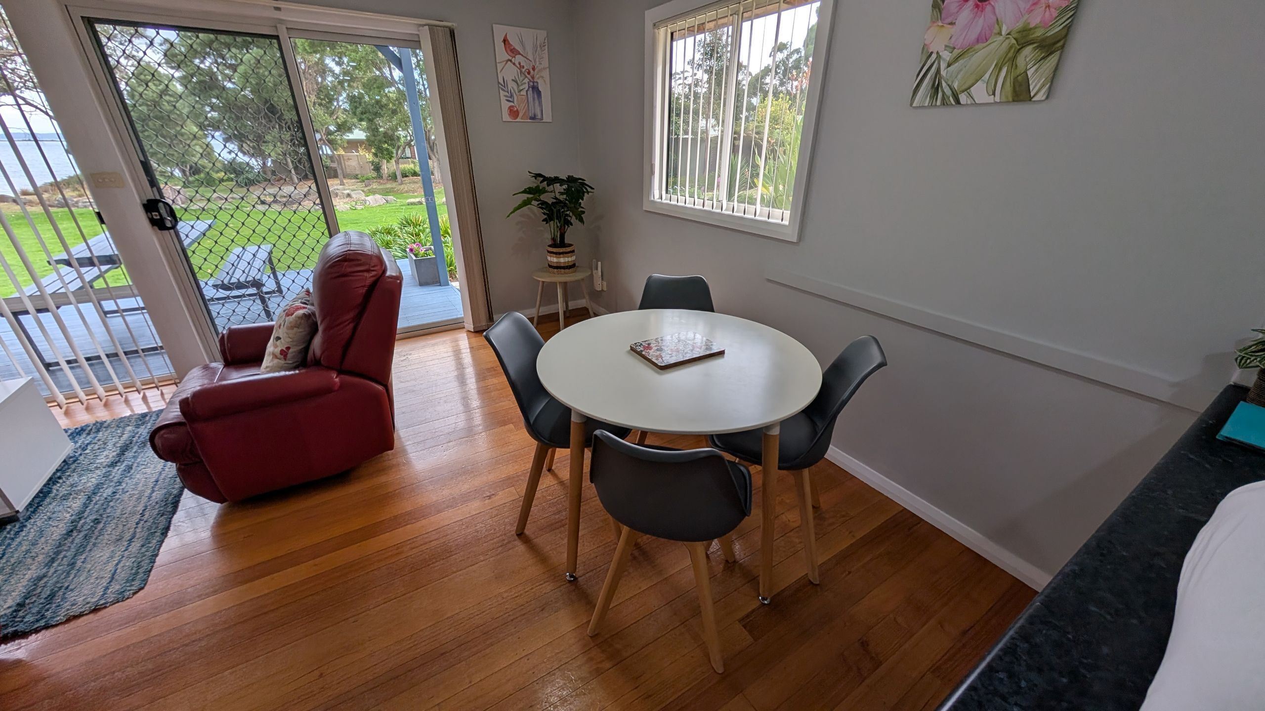 A room with a round table, four chairs, a red sofa, a small potted plant, and a view of a garden through a glass door.