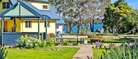 Una casa amarilla con techo azul, una terraza de madera y un jardín con flores y árboles.