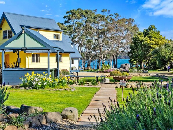 A yellow house with a blue roof, a wooden deck, and a garden with flowers and trees. - Four Seasons Waterfront Villas (Taranna)