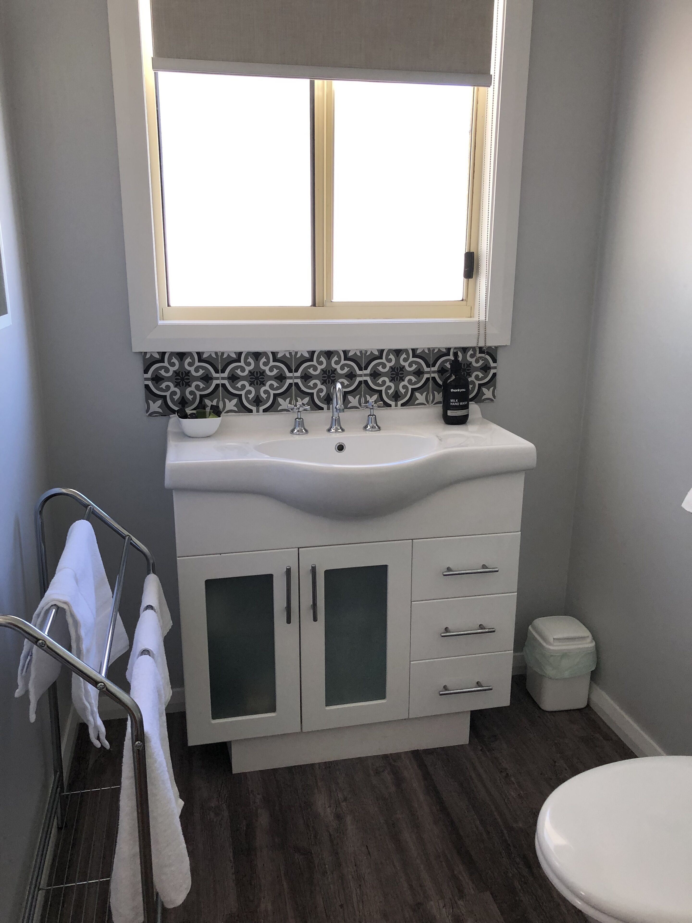 A bathroom with a white sink, two faucets, a mirror, and a window above the sink. There is a towel rack with towels and a trash bin.