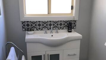 A bathroom with a white sink, two faucets, a mirror, and a window above the sink. There is a towel rack with towels and a trash bin.