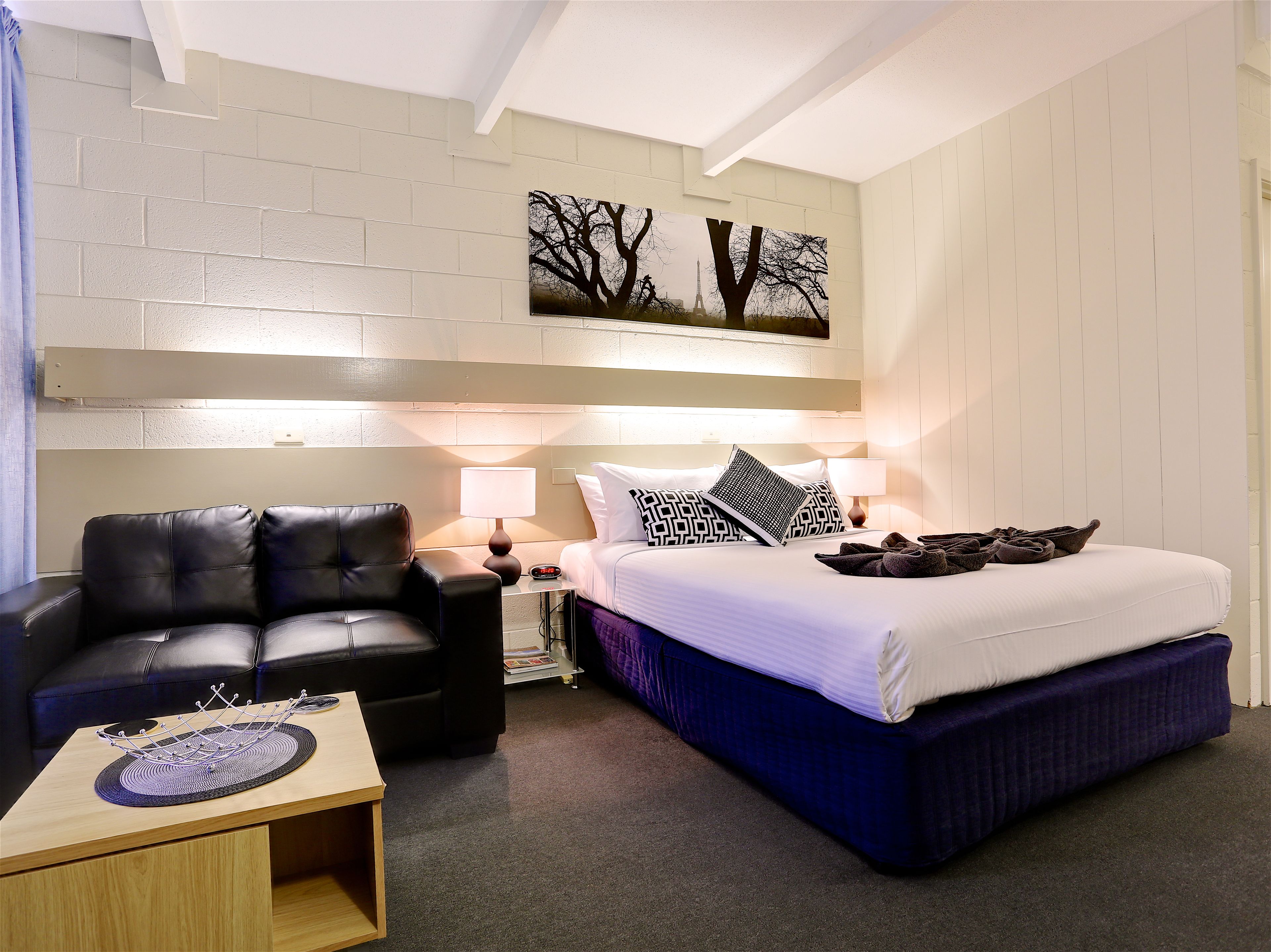 Ein Hotelzimmer mit einem Bett, einem schwarzen Ledersessel, einem kleinen Tisch und einem an der Wand hängenden Kunstwerk.