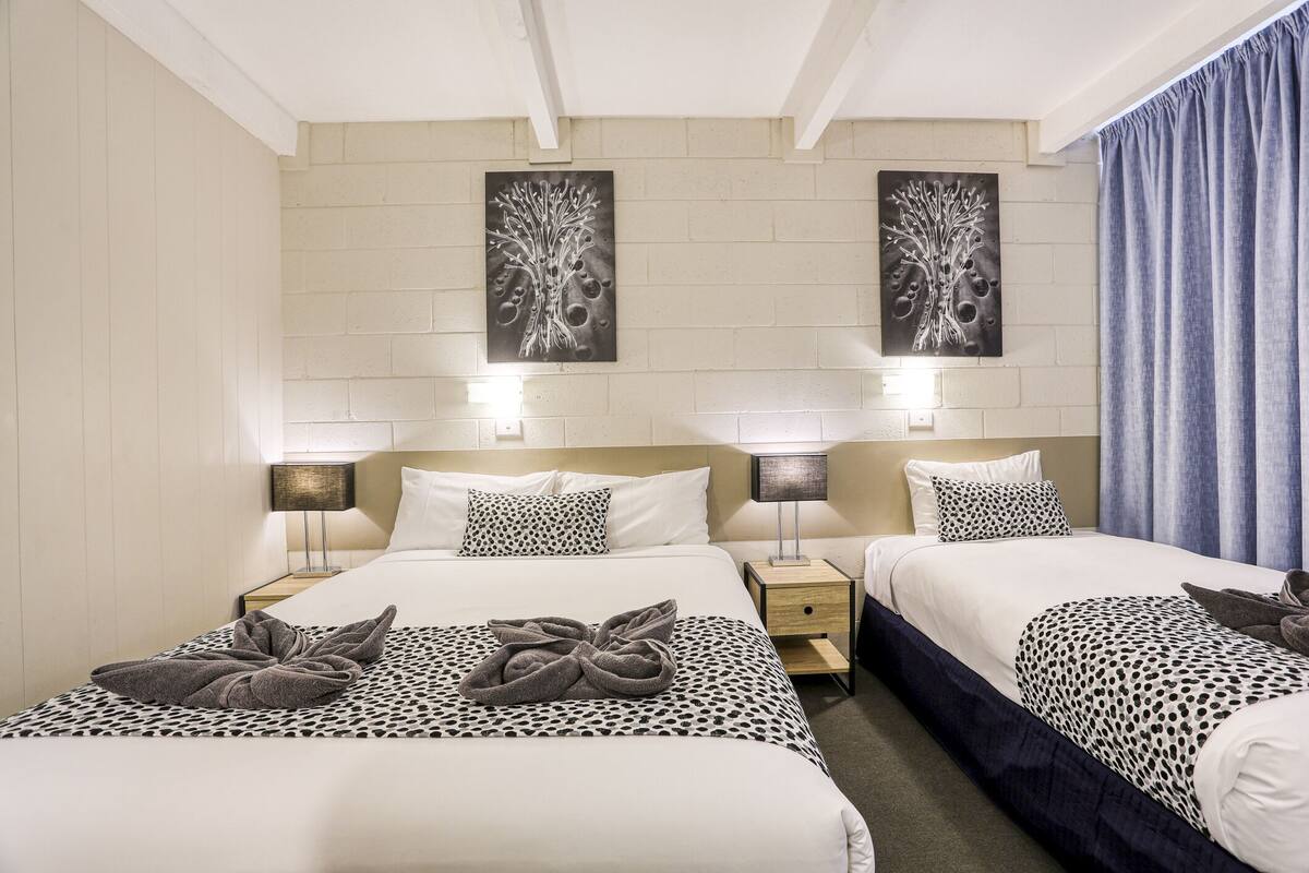 A hotel room with two beds, each with a patterned bedspread and a towel on the bed. There are wall-mounted lamps on either side of the beds, and two wall art pieces above the beds.