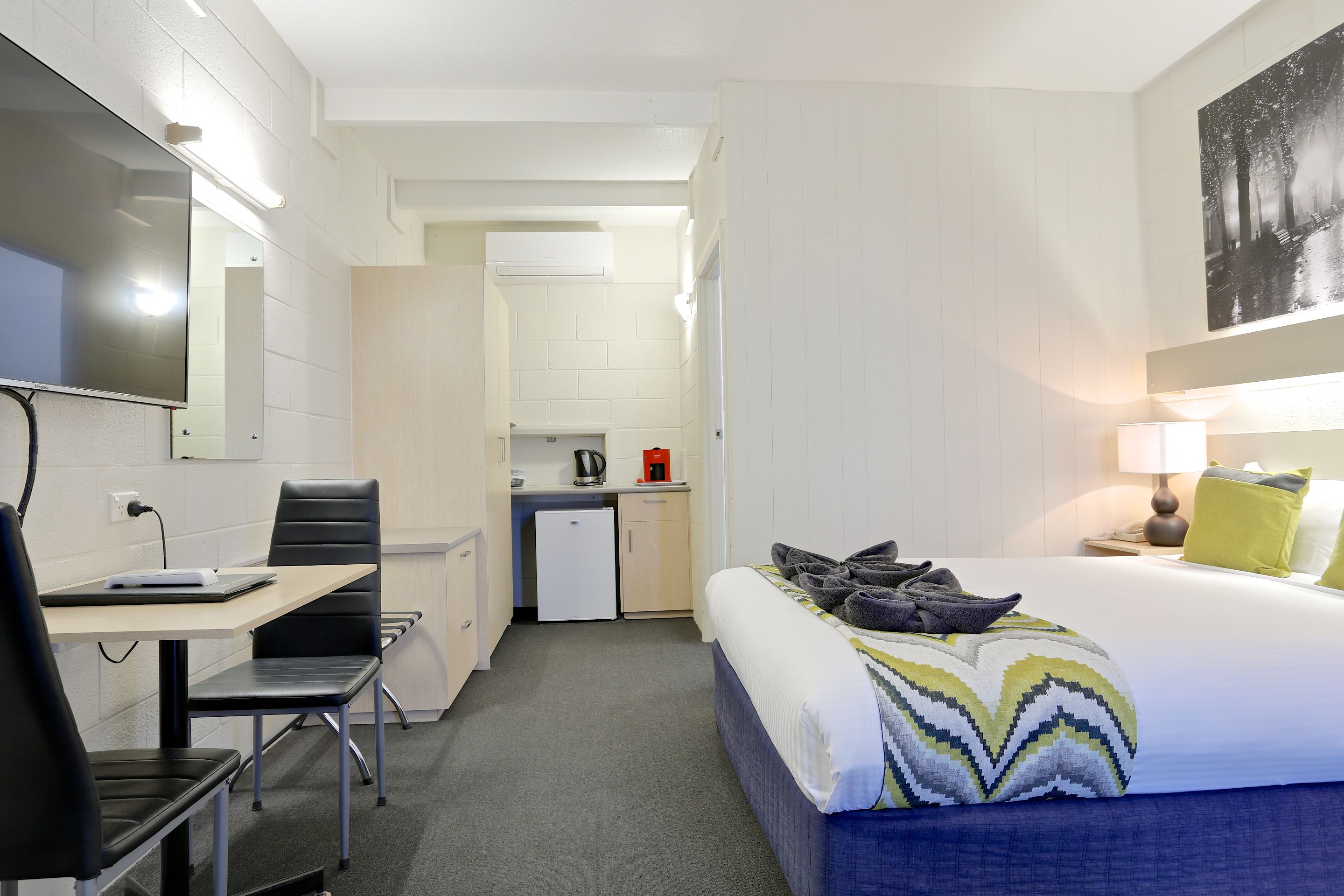 A hotel room with a bed, a desk, a chair, a kitchenette, and a mirror.