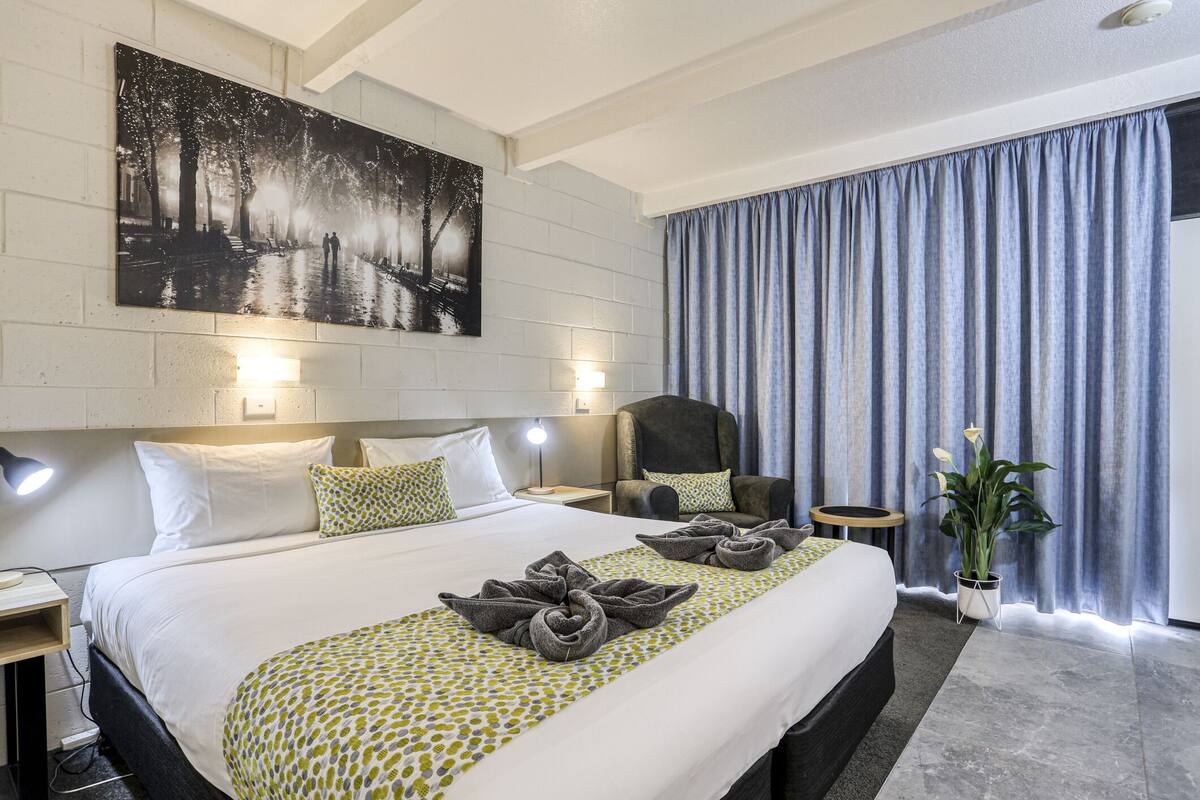 A hotel room with a large bed, a chair, a nightstand, a lamp, a plant, and a painting on the wall.