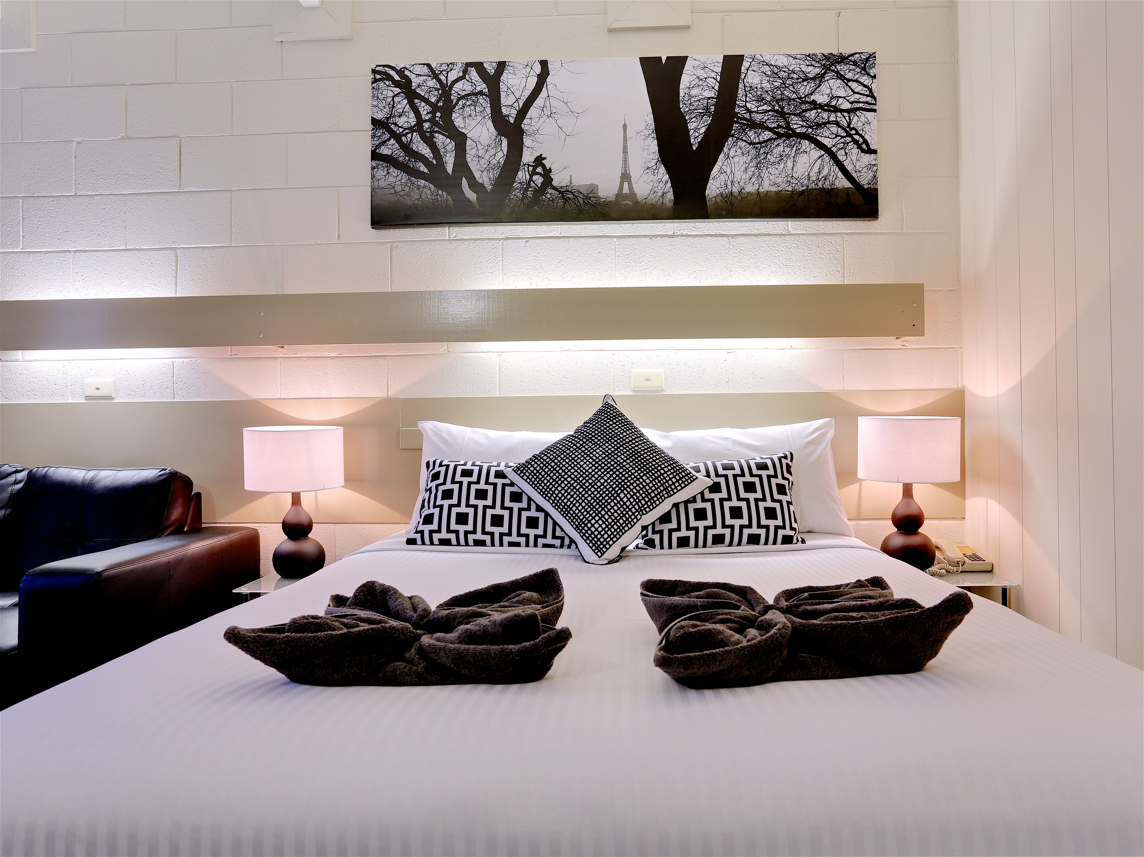 Habitación de hotel con cama, dos almohadas, manta, mesita de noche, lámpara, teléfono y un cuadro colgado en la pared.