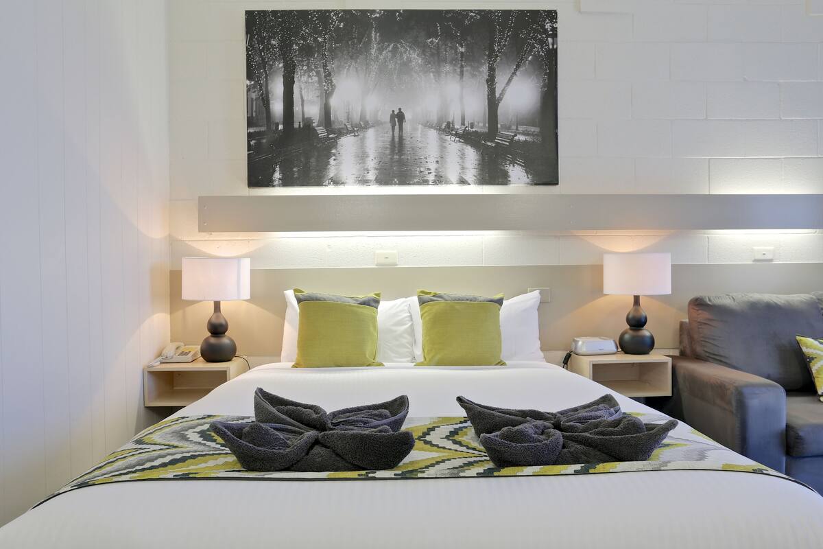A hotel room with a large bed, two bedside tables with lamps, a grey sofa, and a wall-mounted artwork of a forest path.