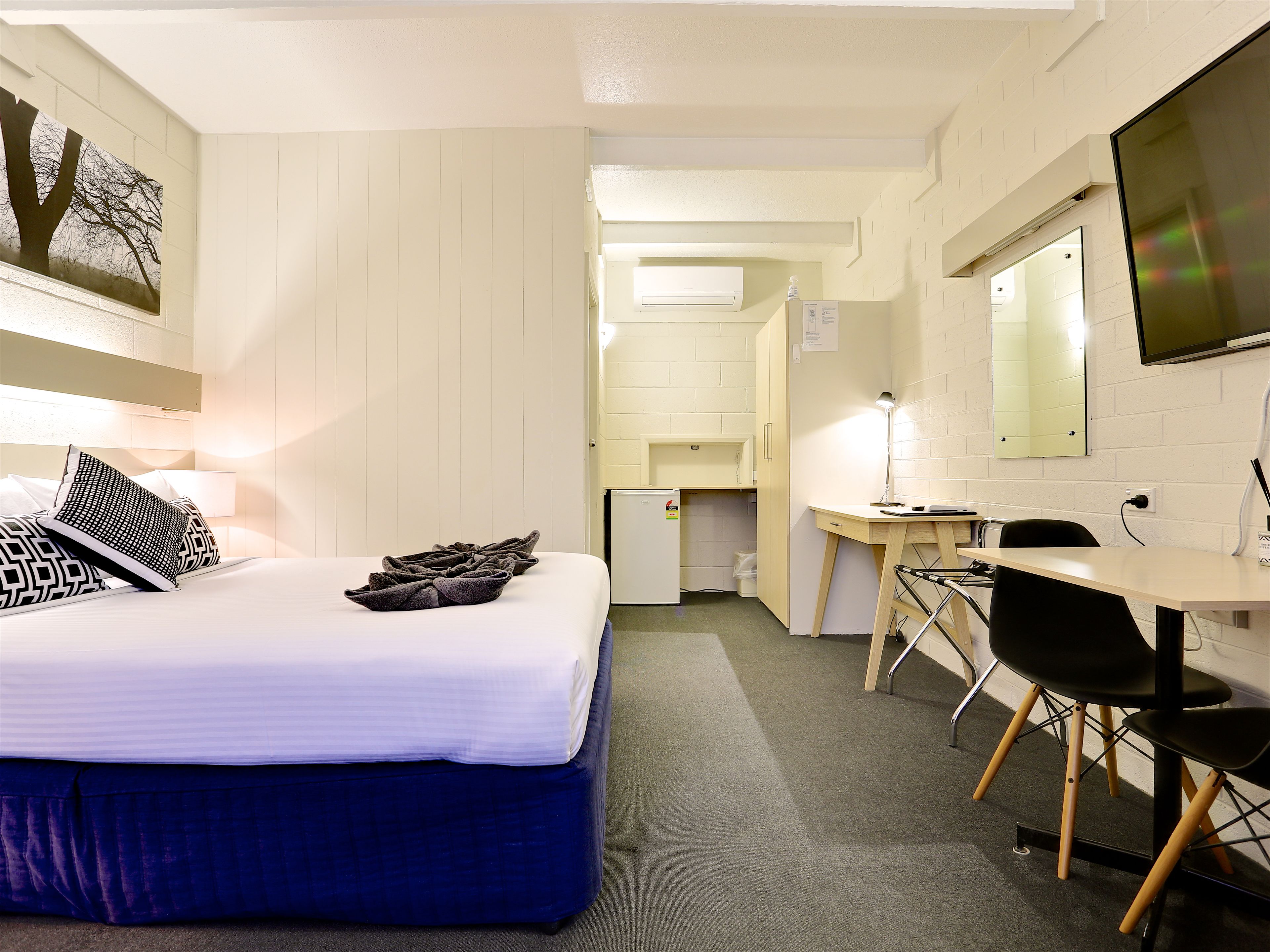 A hotel room with a bed, a desk, a chair, a mirror, and a small kitchenette.