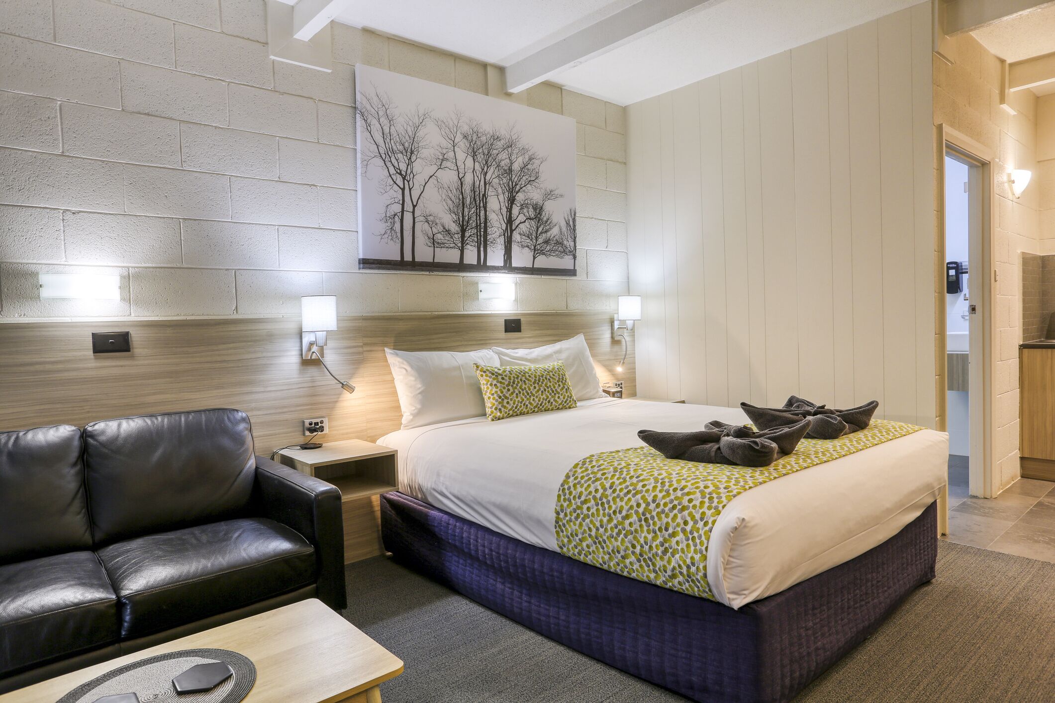 A hotel room with a bed, a black leather sofa, a wooden nightstand, and a wall-mounted artwork.