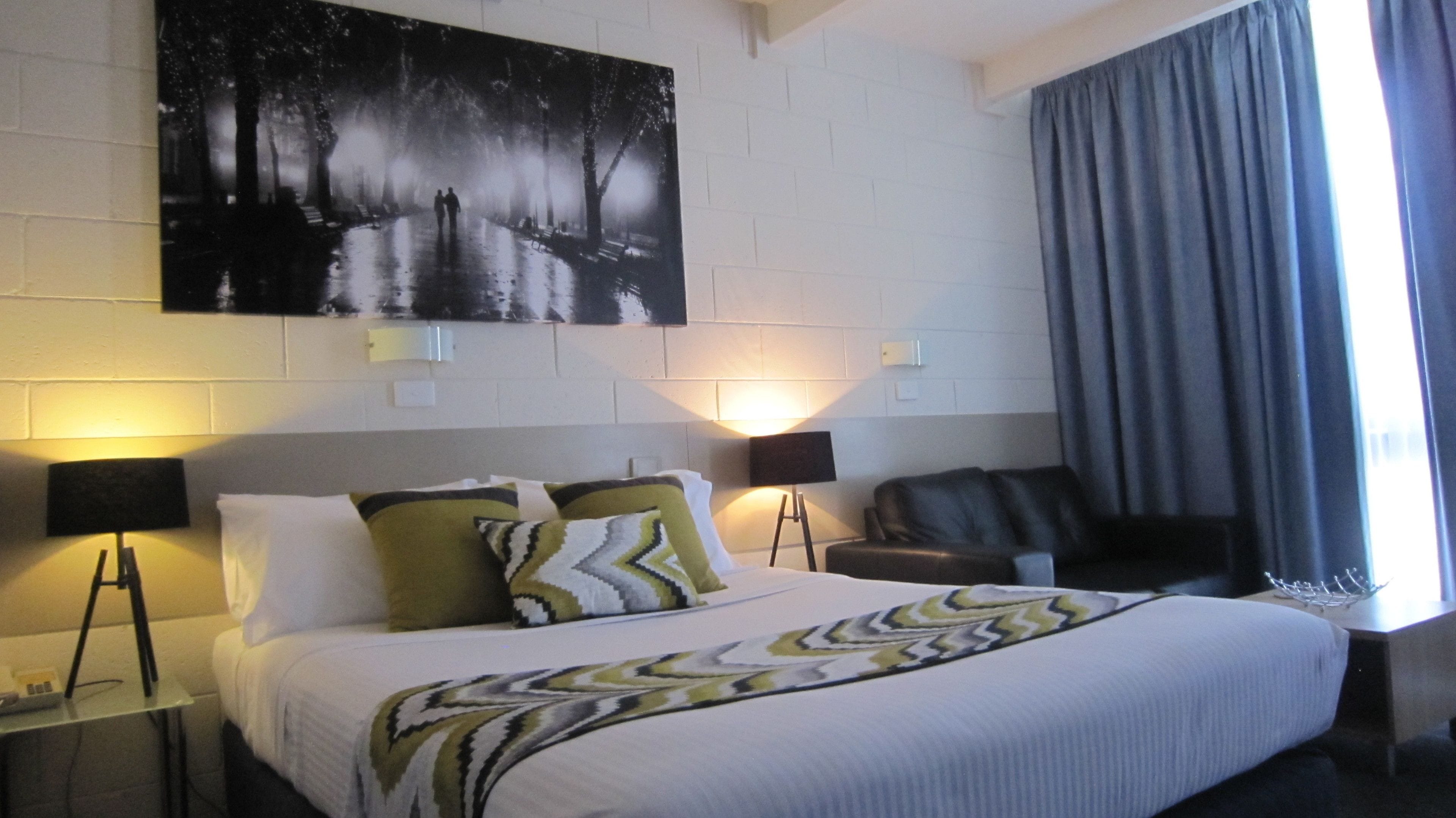 A hotel room with a bed, two bedside lamps, a chair, a painting on the wall, and a window with curtains.