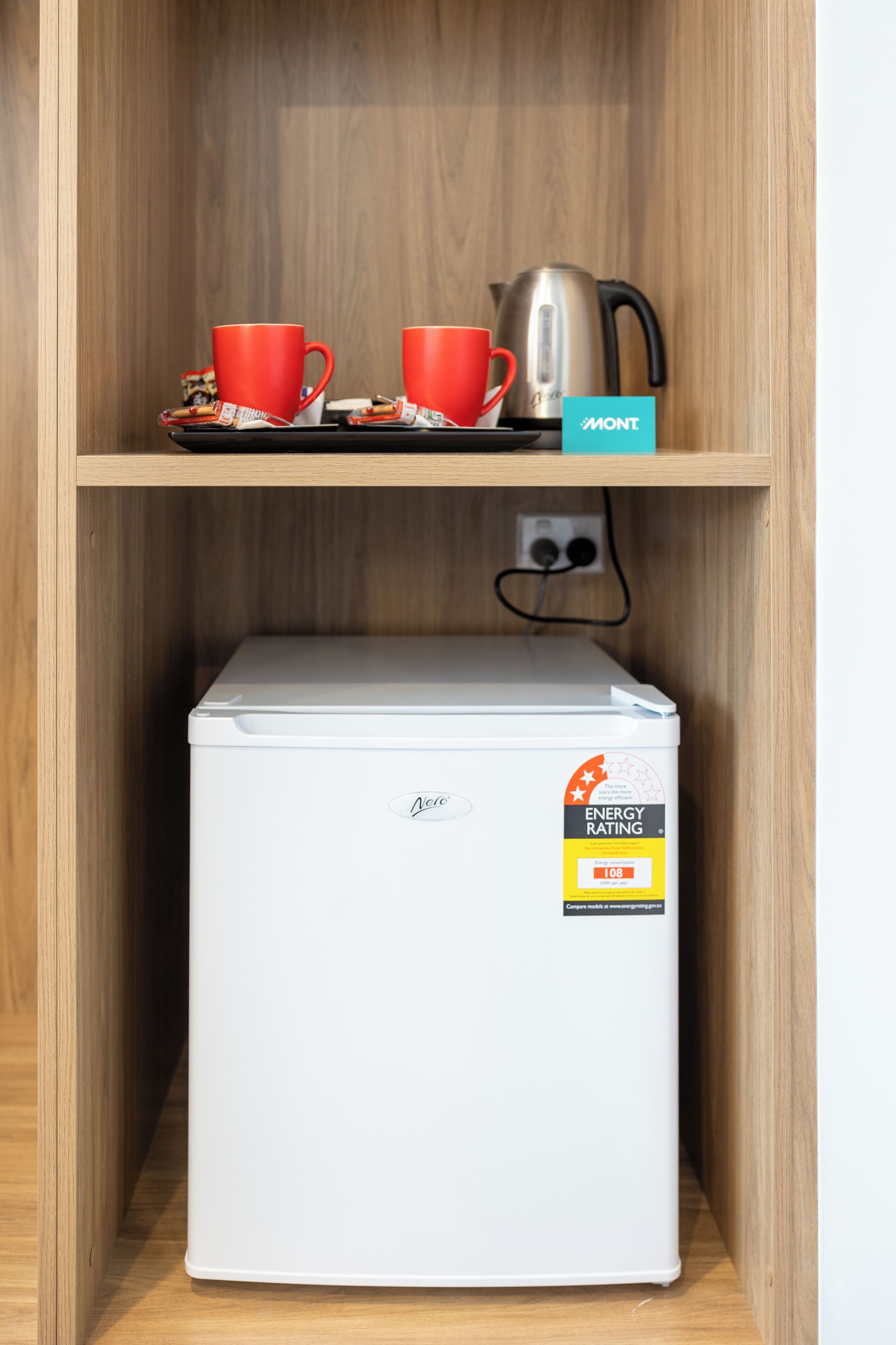 A compact white mini-fridge with an energy rating sticker, two red cups on a tray, and a kettle on a shelf above.