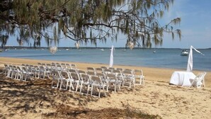 A beach wedding setup with white chairs and tables under a tree, overlooking the sea with boats. - Coochiemudlo Island Seminara Apartments (Coochiemudlo Island)
