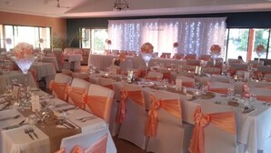 A banquet hall with tables set for dining, chairs with orange sashes, and a large floral centerpiece. - Coochiemudlo Island Seminara Apartments (Coochiemudlo Island)