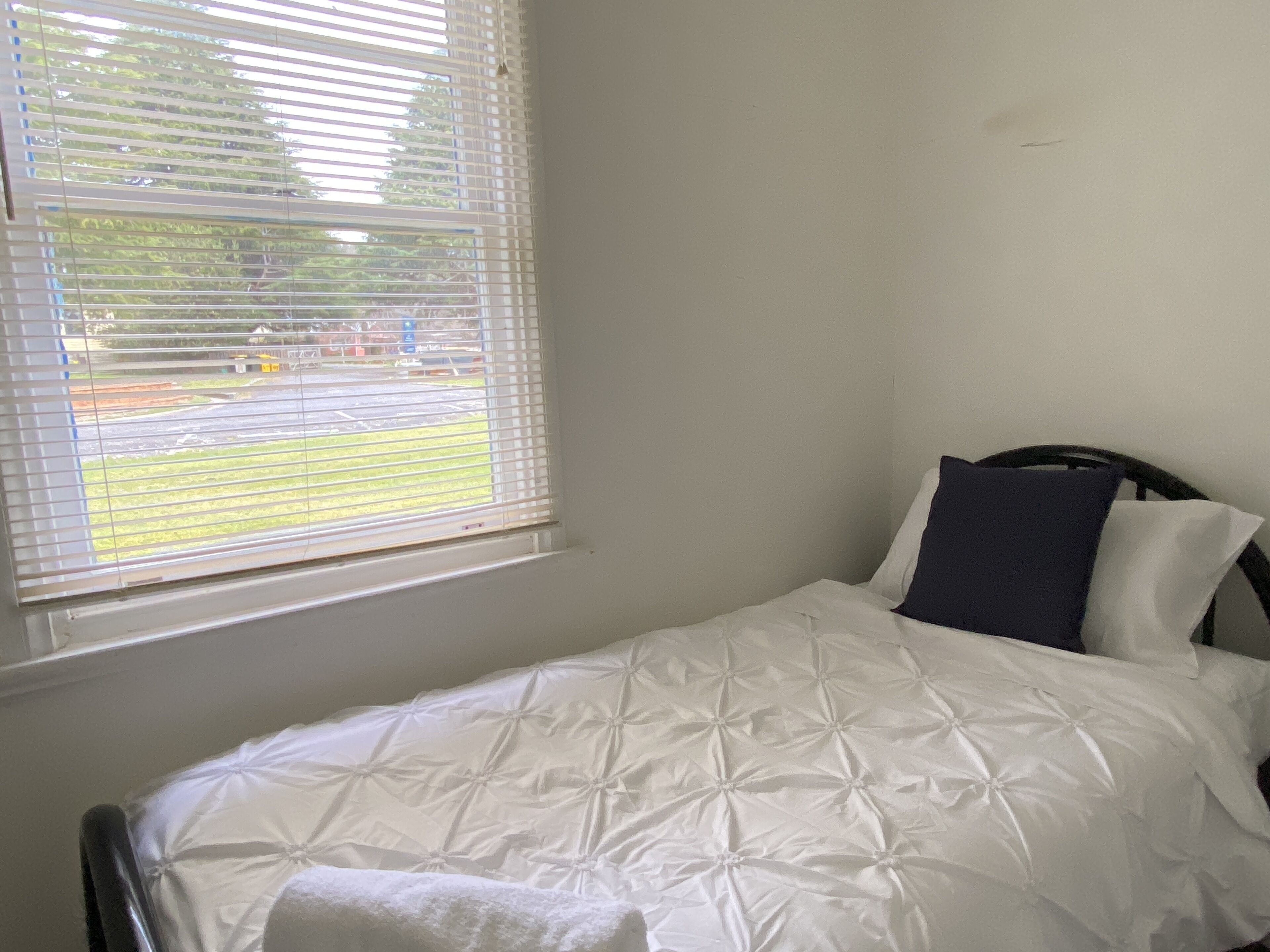 A bedroom with a bed, a window with blinds, and a view of a parking lot and trees.