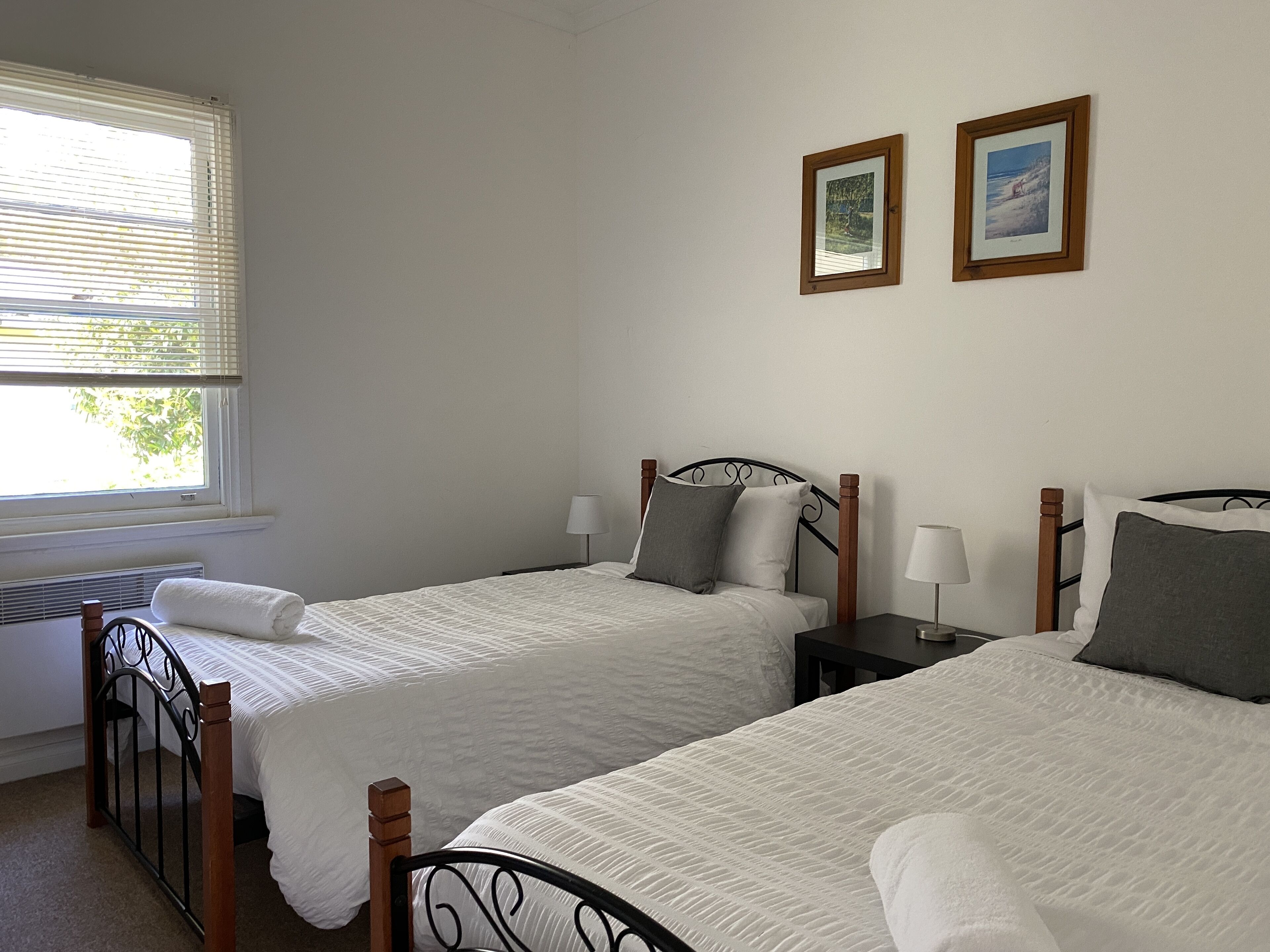 A bedroom with two single beds, a window with blinds, a radiator, and two framed pictures on the wall.