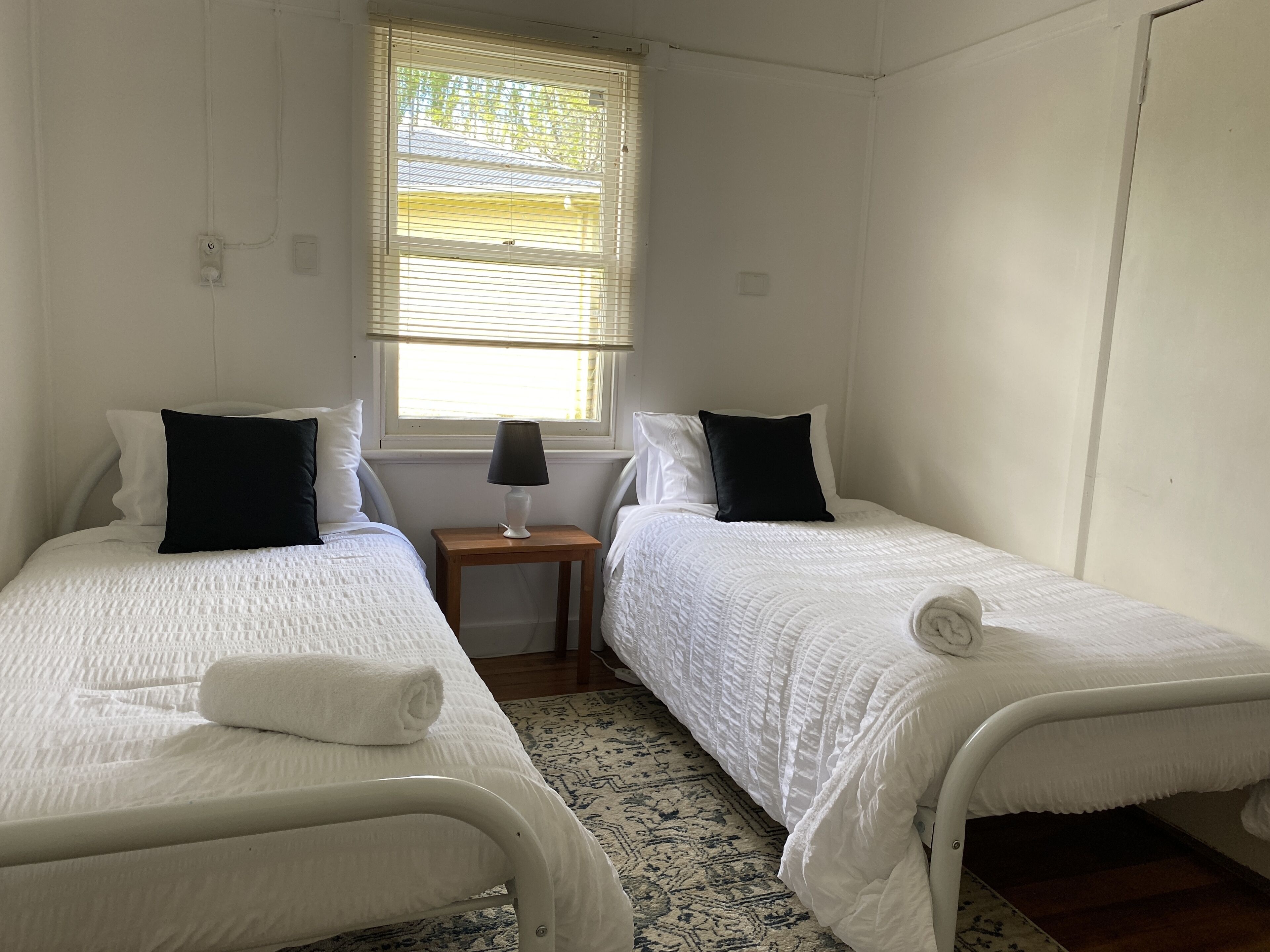 A twin room with two beds, a small table with a lamp, and a window with blinds.