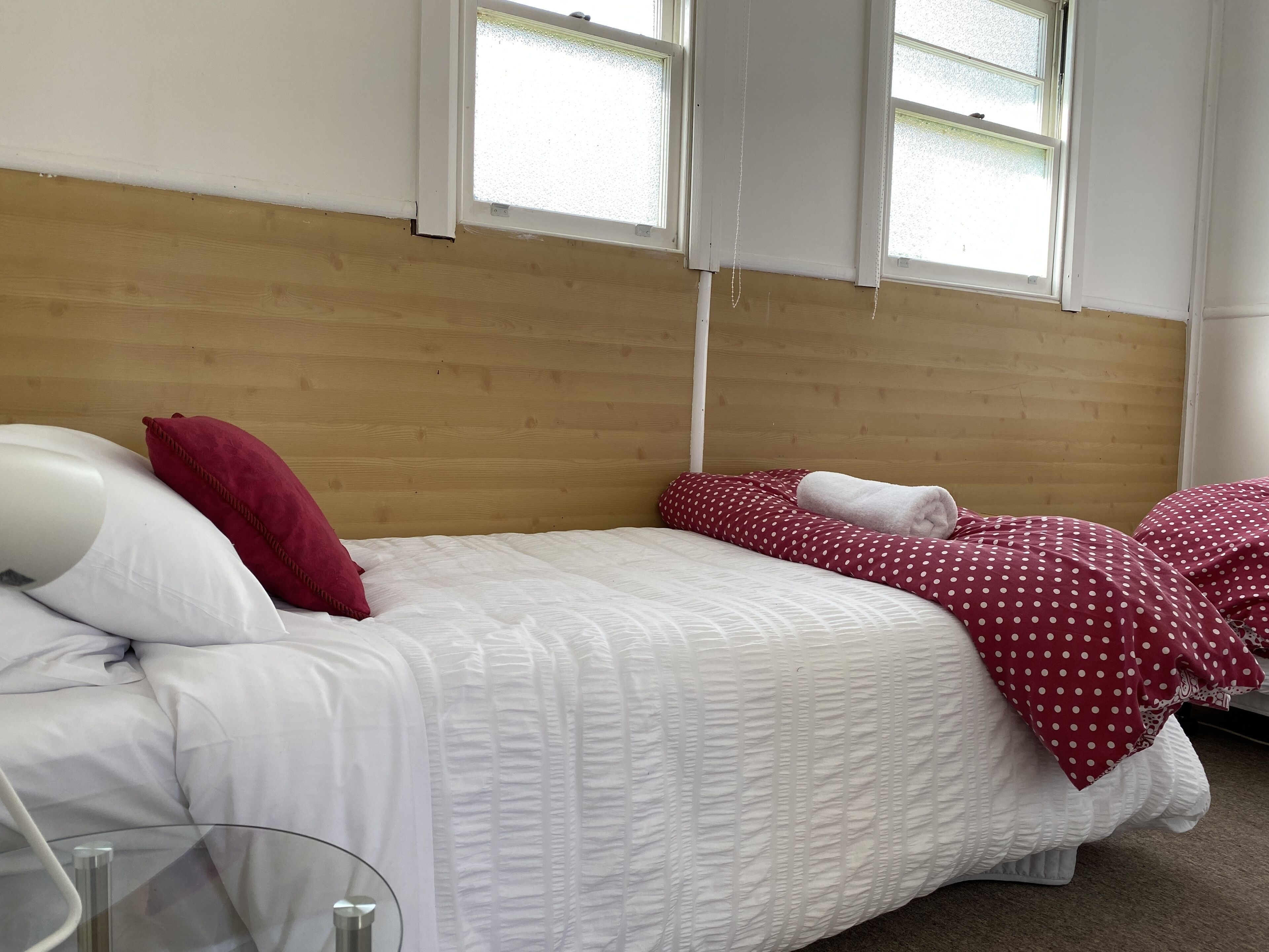 Een enkel bed met witte beddengoed en rode stippelhoeden, een nachtkastje met een lamp, en een raam met zonwering.
