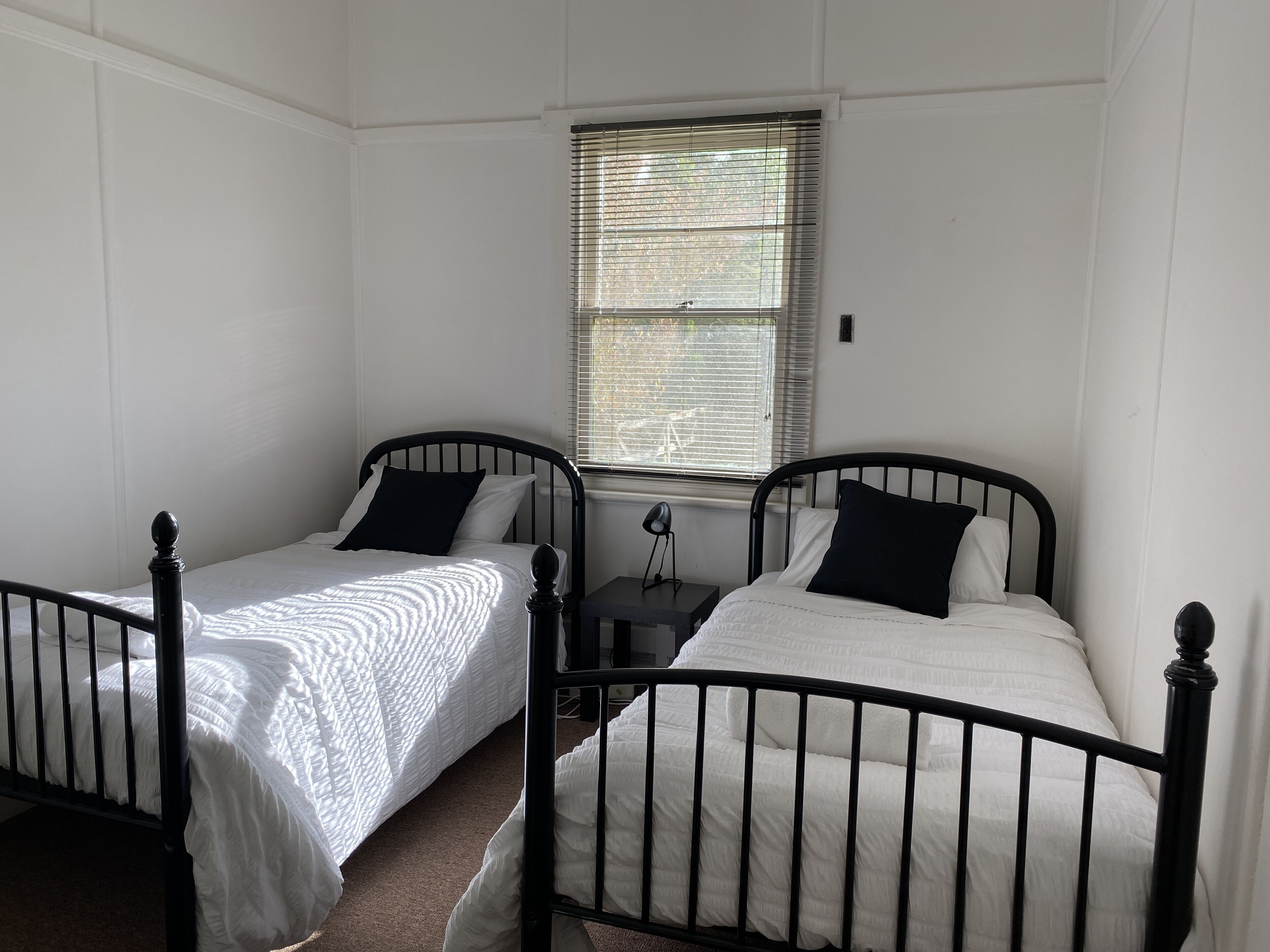 Habitación con dos camas individuales, cada una con cabecero negro y ropa de cama blanca. Se aprecia una ventana con persianas, una mesita de noche con lámpara, y un interruptor de luz en la pared.