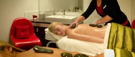 A massage therapy session with a client lying on a massage table, a therapist applying stones to the client's back, and a red chair in the background.