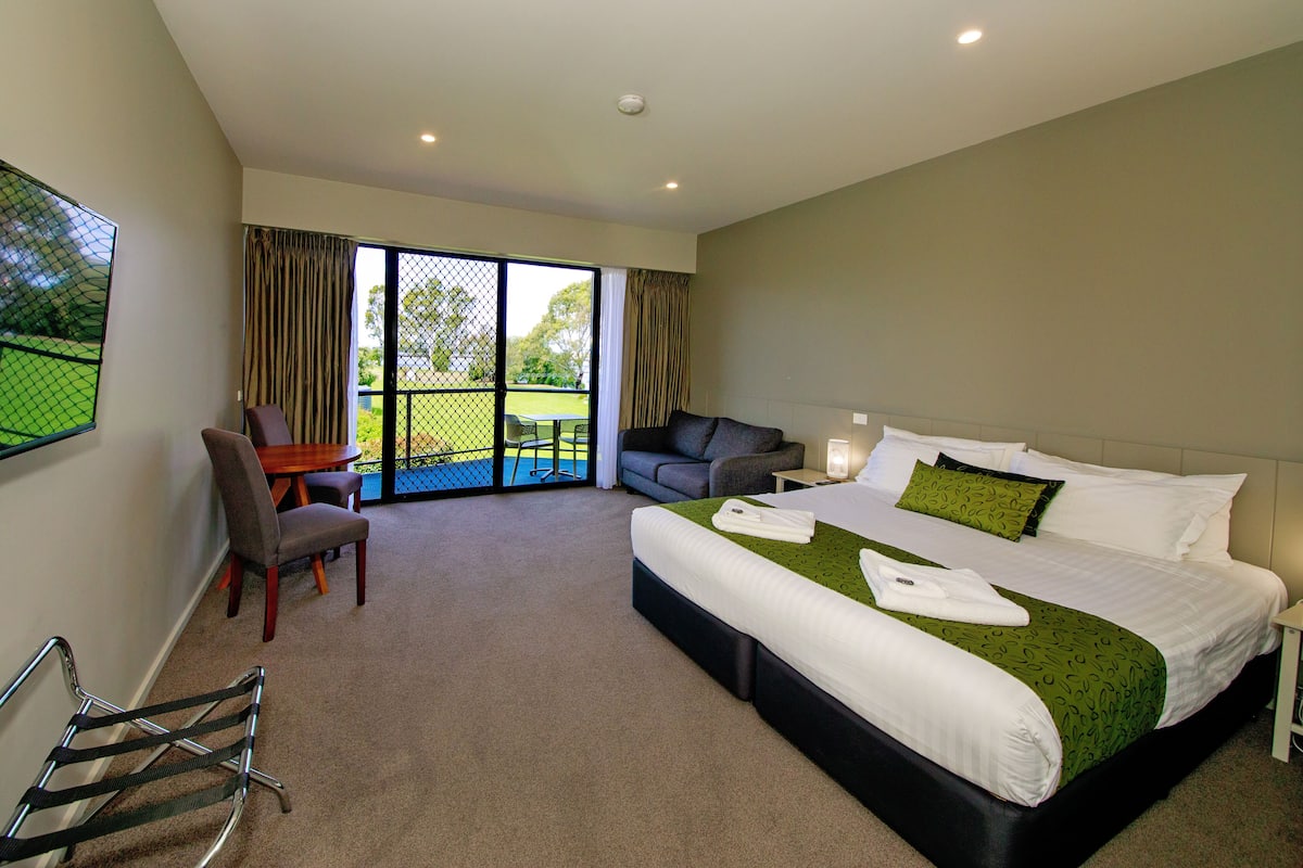 A hotel room with a large bed, a desk, a chair, a sofa, and a balcony with a view of the outdoors.