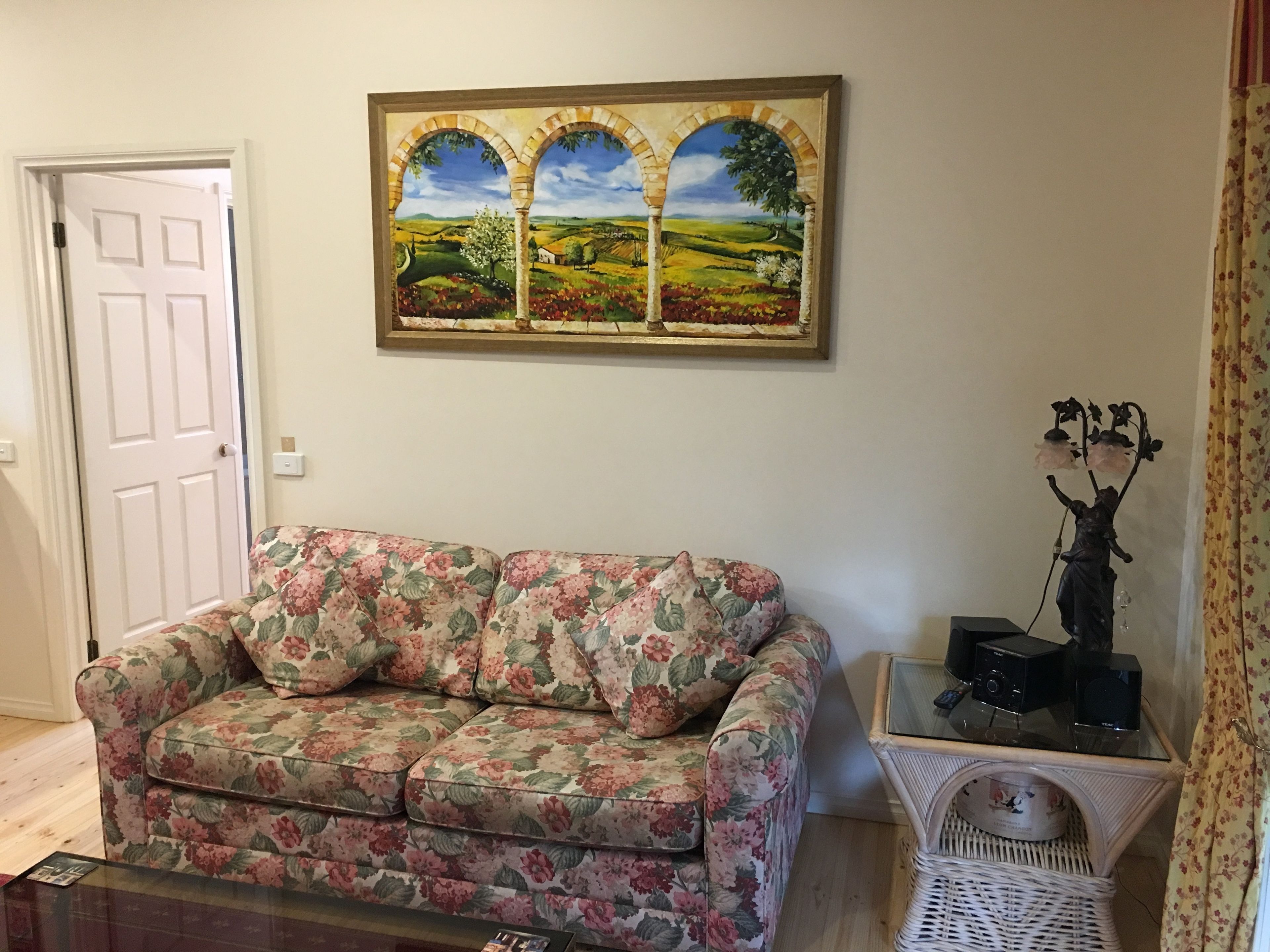 A living room with a floral-patterned sofa, a painting of a landscape, a small table with a statue, and a wicker basket.