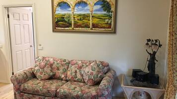 A living room with a floral-patterned sofa, a painting of a landscape, a small table with a statue, and a wicker basket.