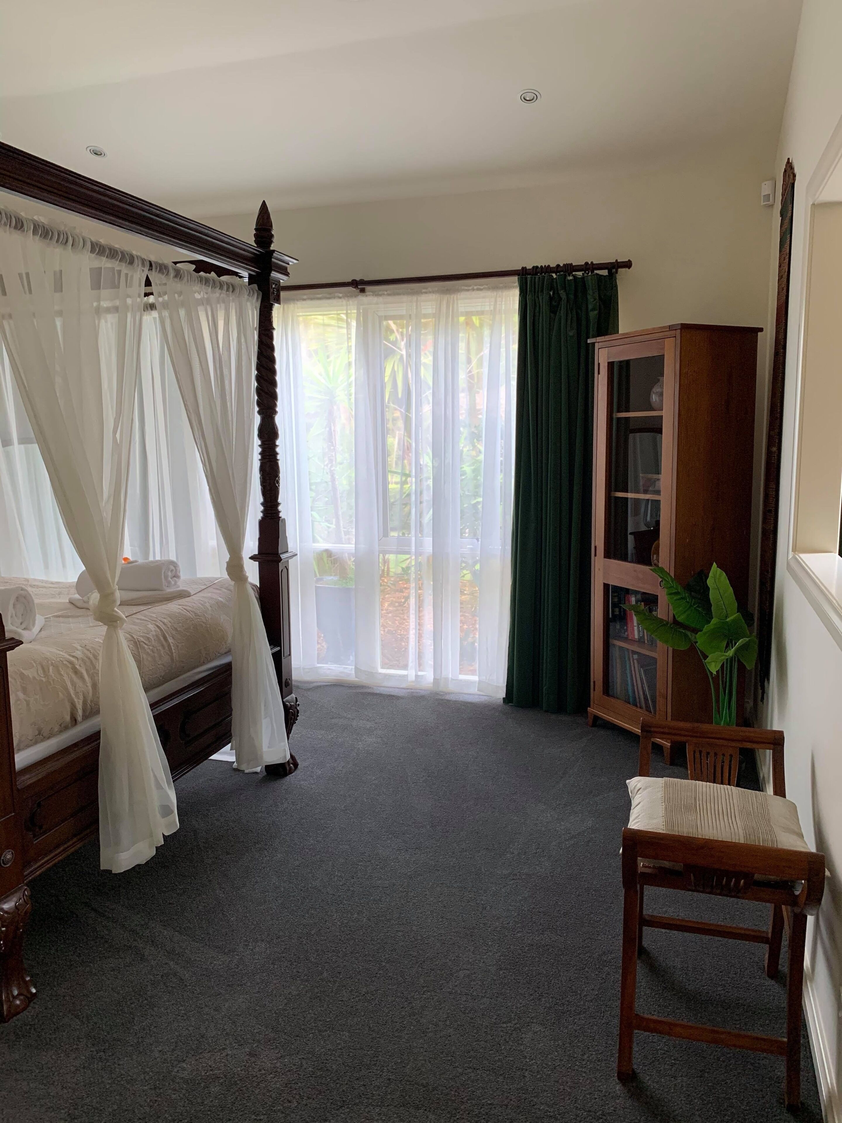 A bedroom with a four-poster bed, a wooden cabinet, a chair, and a large window with sheer curtains.