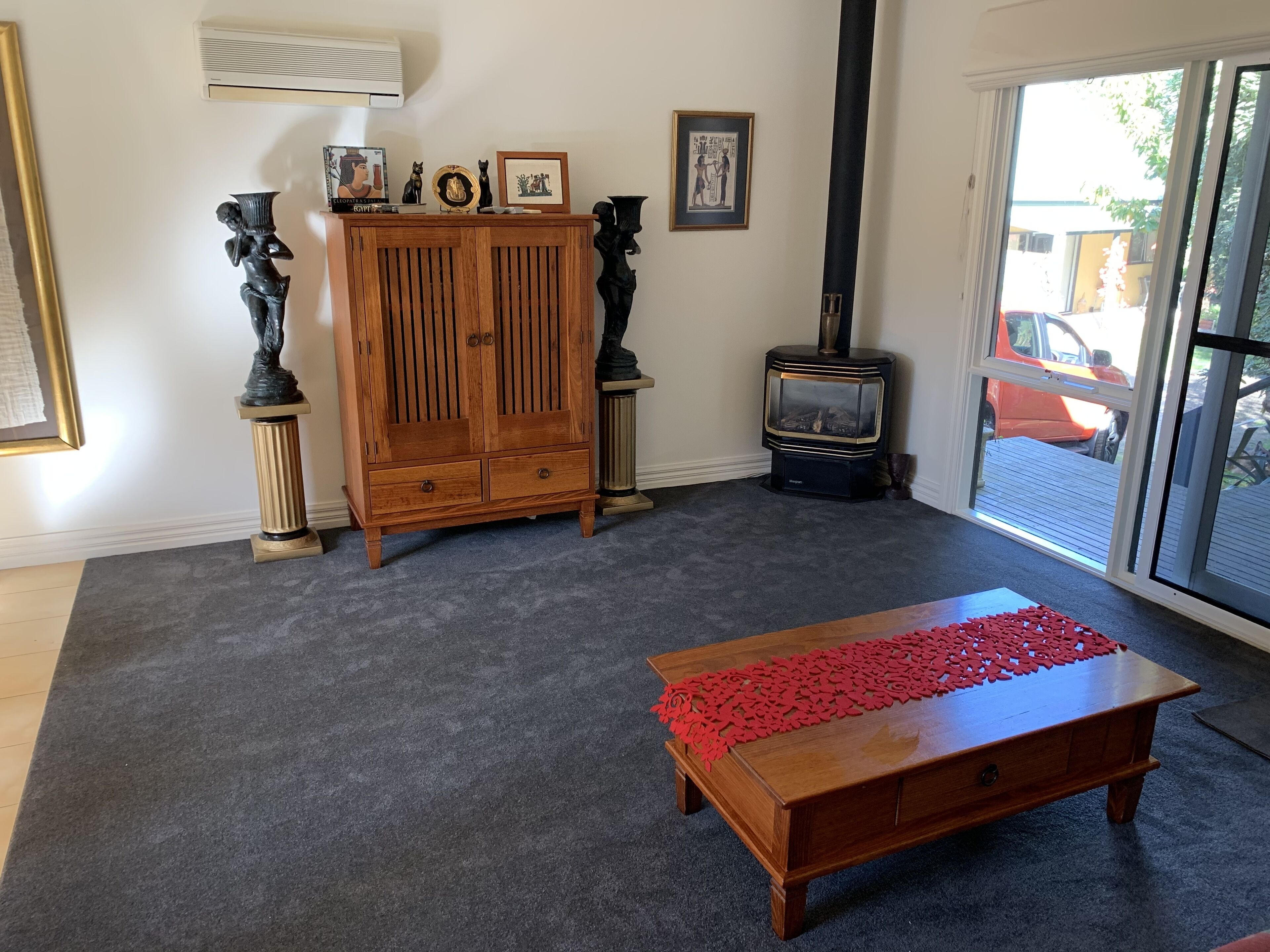 A living room with a wooden cabinet, a red decorative runner on a coffee table, a fireplace, and a large window with a view of the outside.