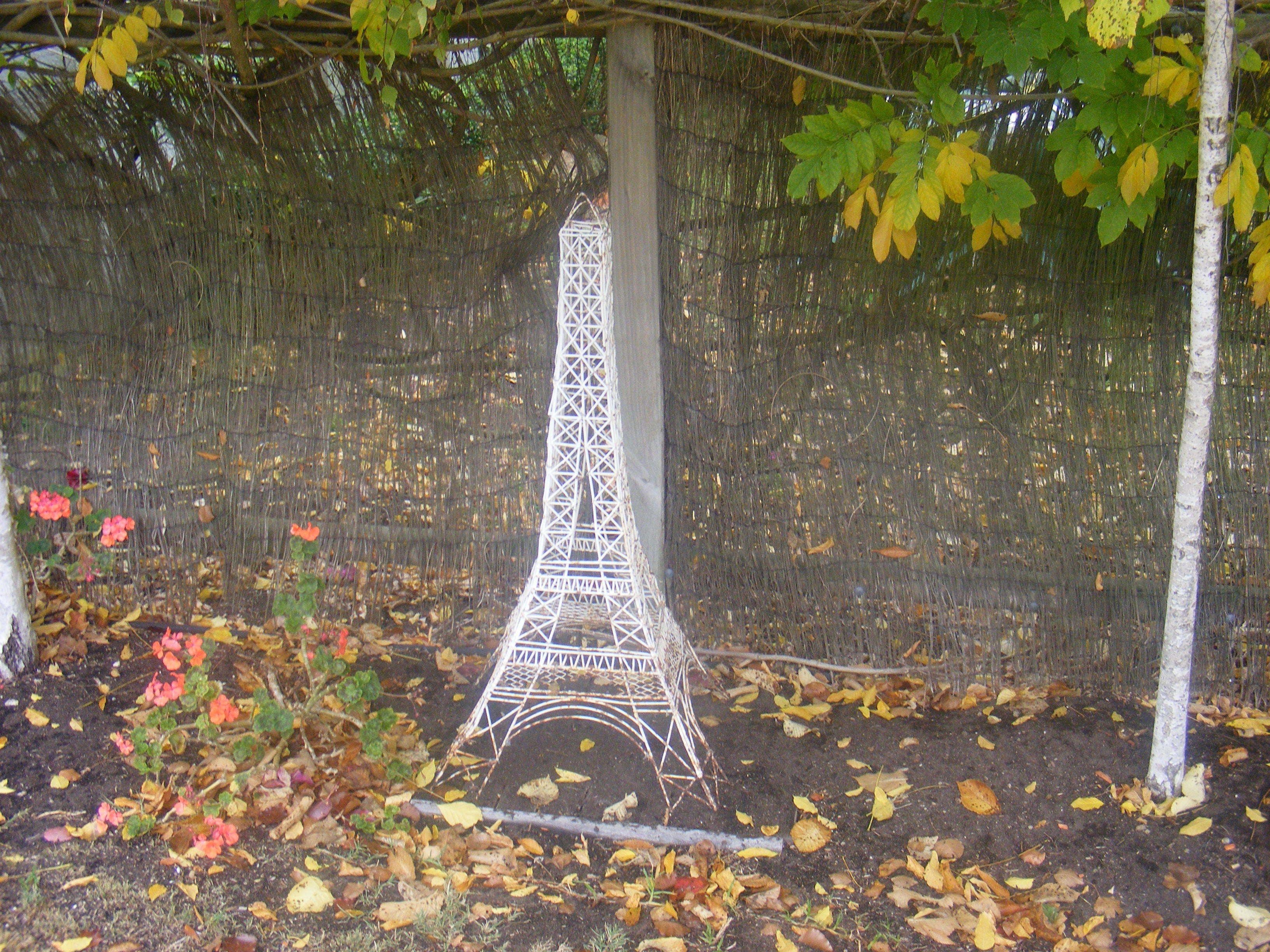 A model of the Eiffel Tower in a garden setting with leaf-covered ground and a mesh fence.