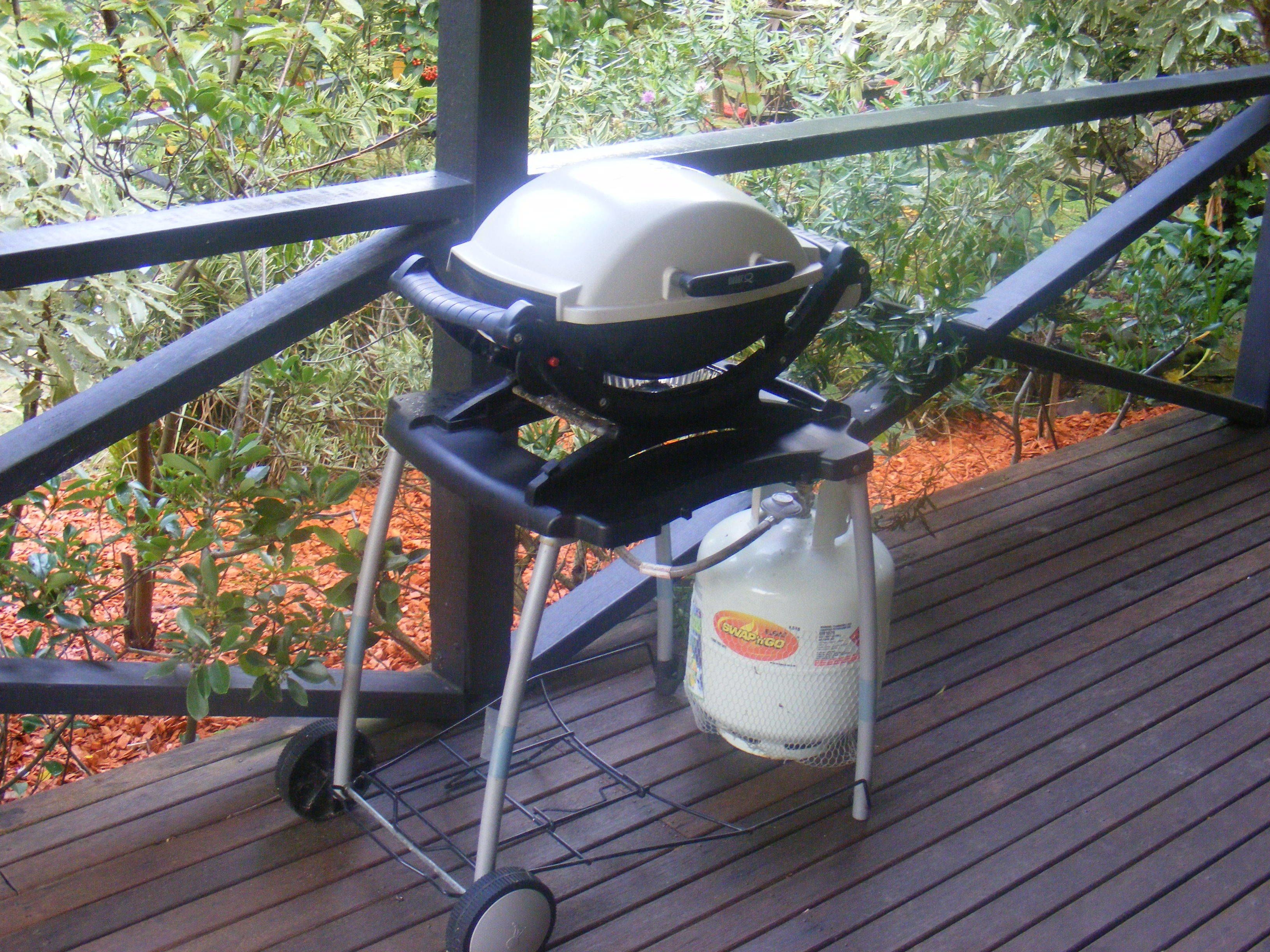 A barbecue grill on a wooden deck with a metal railing, surrounded by greenery.