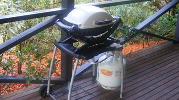 A barbecue grill on a wooden deck with a metal railing, surrounded by greenery.