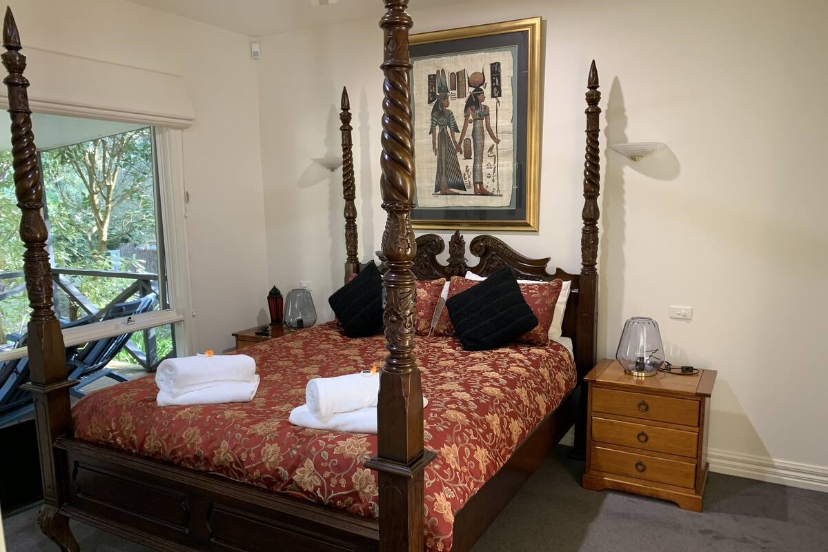 A four-poster bed with a red floral bedspread, two black pillows, and a nightstand with a lamp.