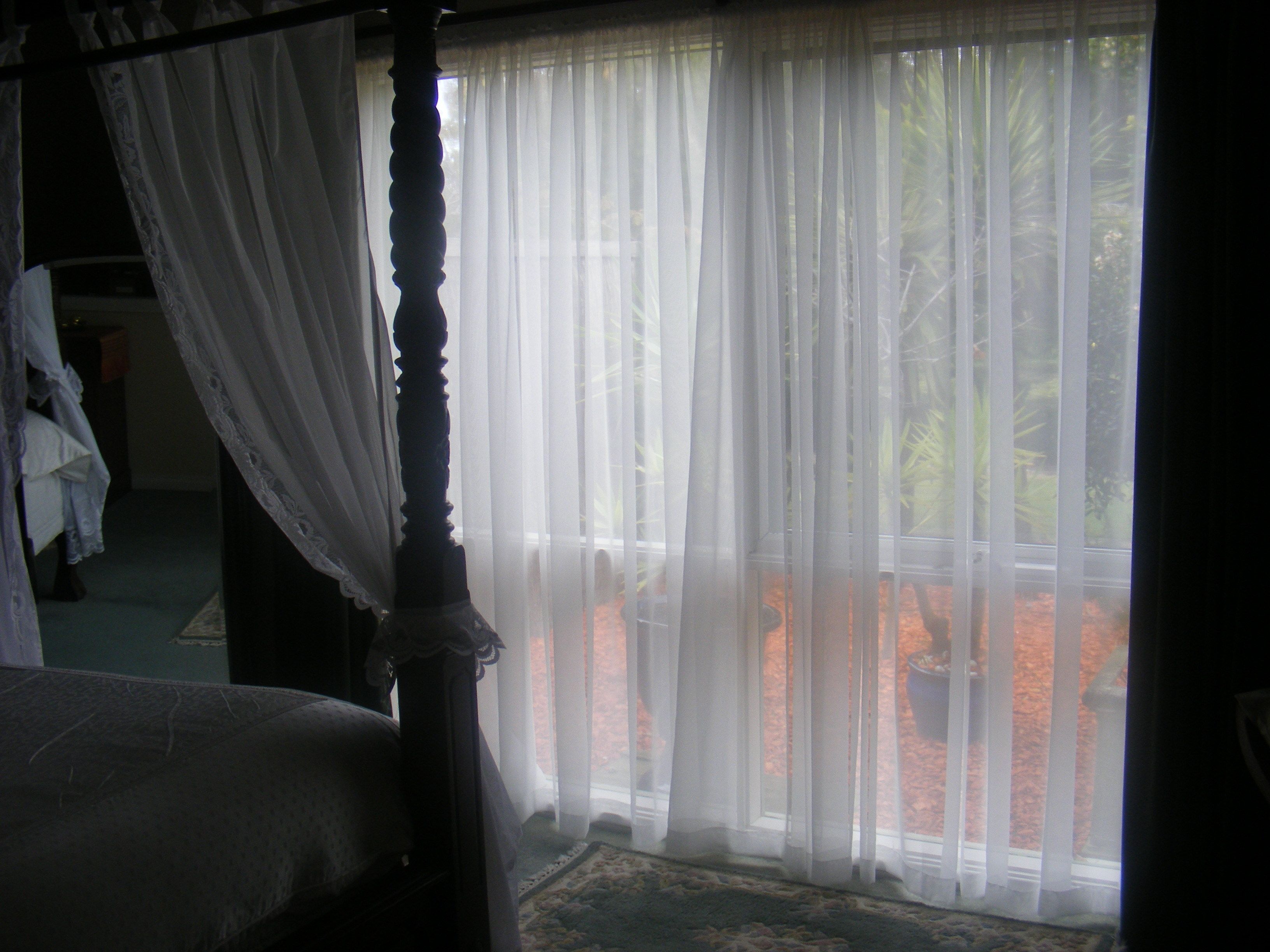 A bedroom with a four-poster bed, sheer curtains, and a view of a landscaped outdoor area.