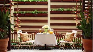 An outdoor seating area with a table set for two, surrounded by wooden chairs, with a backdrop of vertical wooden slats and greenery.