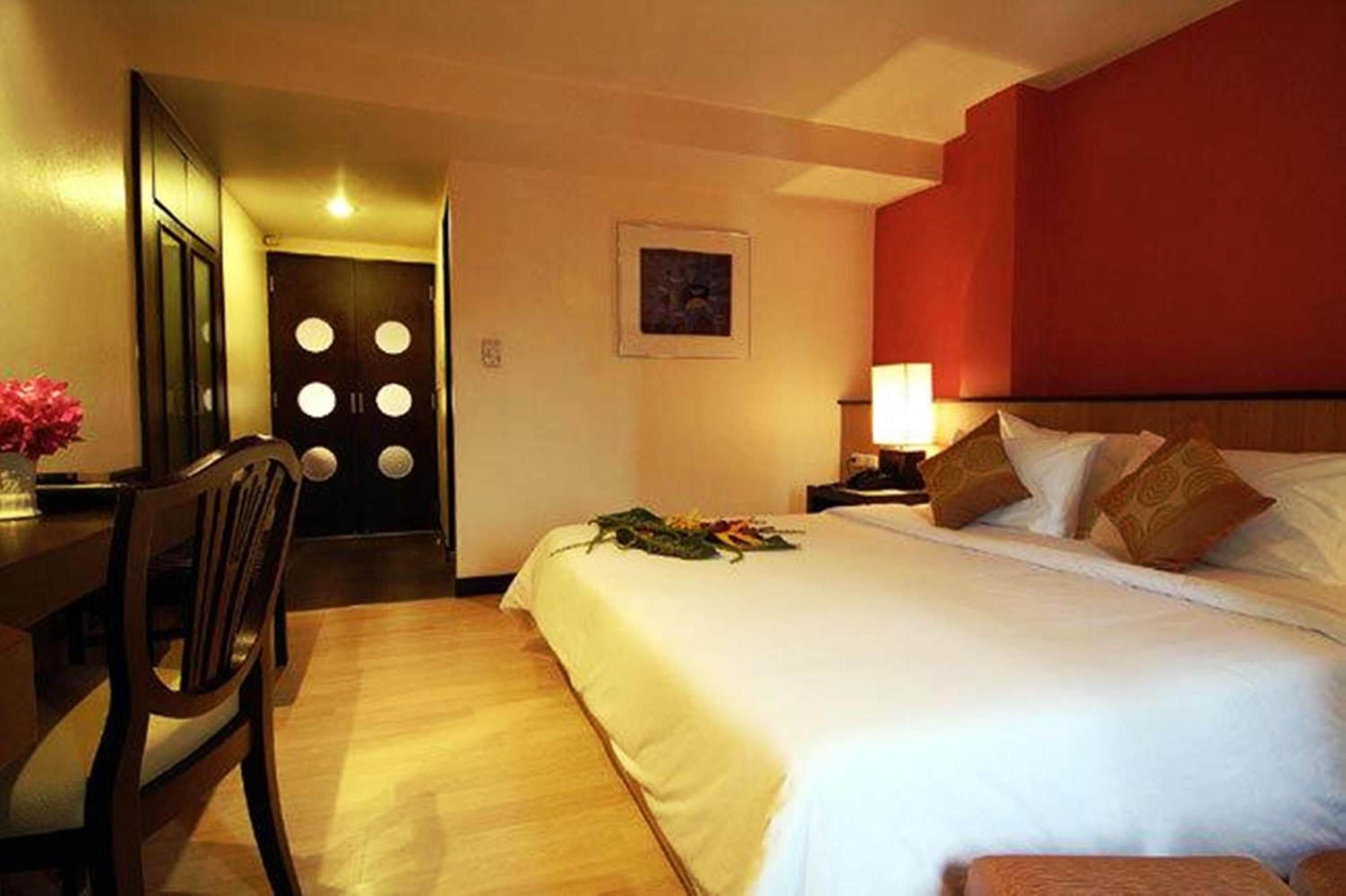 A hotel room with a bed, a desk with a chair, a nightstand with a lamp, and a door with circular windows.