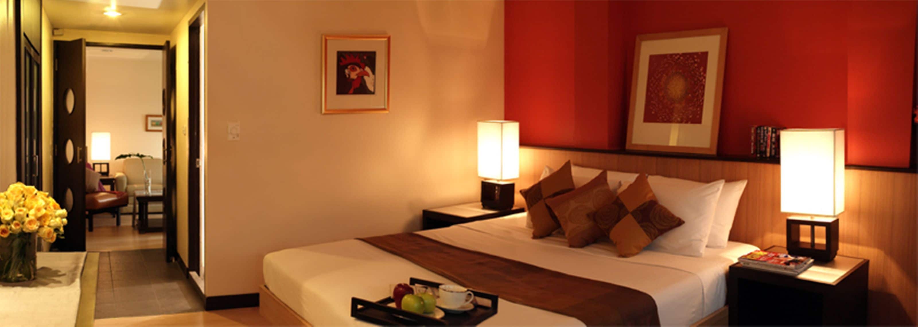 Une chambre d’hôtel avec un grand lit, deux lampes de chevet, un plateau avec des fruits et une tasse, et un mur rouge orné de tableaux.