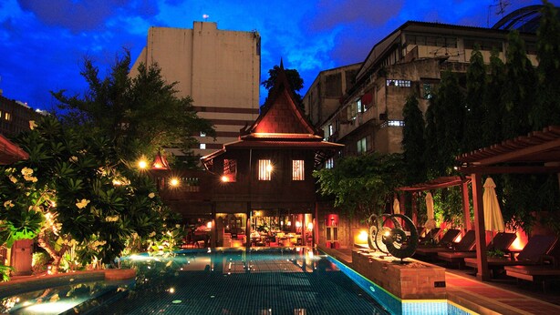Una piscina con un edificio tradicional al fondo, rodeada de abundante vegetación y zonas de estar al aire libre.