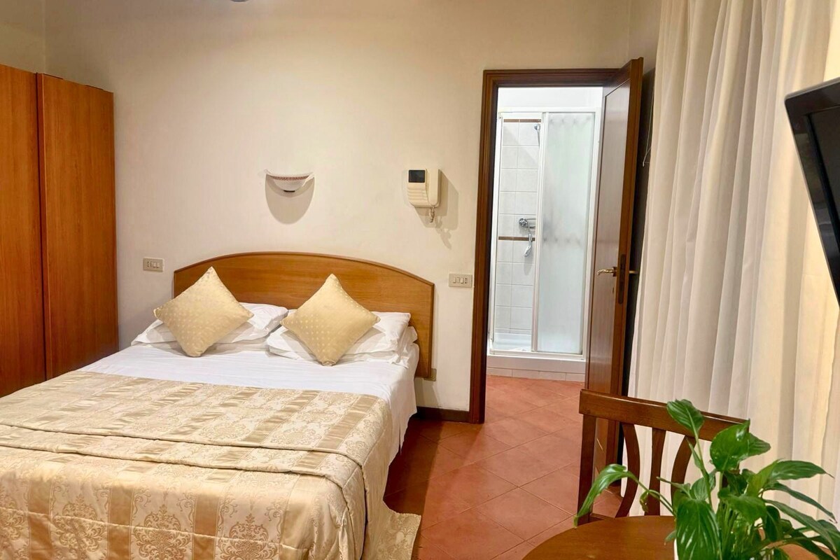 Un dormitorio con cama, un armario de madera, una mesita de noche con una planta y un baño con ducha.