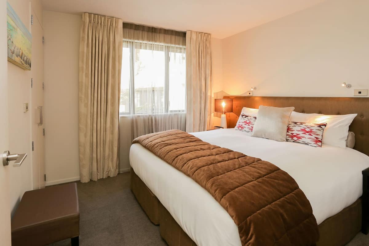 A hotel room with a large bed, a bedside table, a window with curtains, and a wall-mounted lamp.
