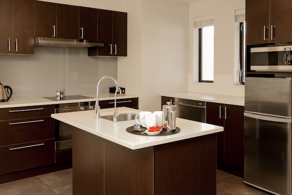 A modern kitchen with dark wooden cabinets, stainless steel appliances, and a white countertop island. - Village Lake Apartments (Hanmer Springs)