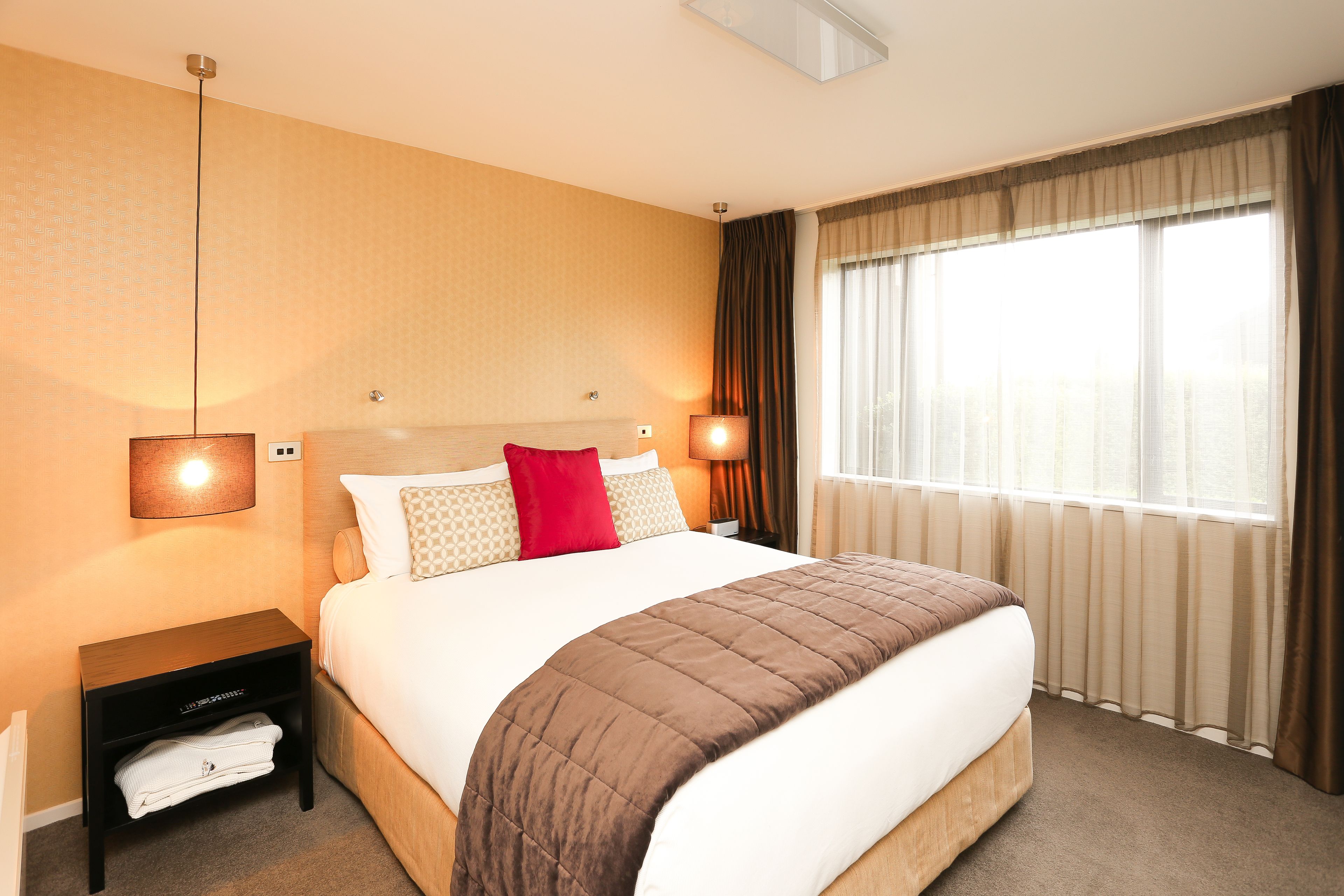 A neatly made bed with a brown quilt, white linens, and a red pillow. There are two bedside lamps, a nightstand with a folded towel, and a window with curtains.