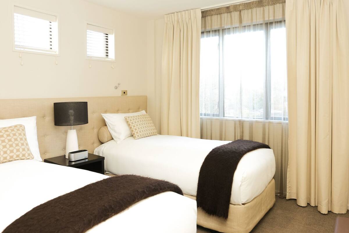 A hotel room with two beds, a nightstand with a lamp, a window with curtains, and a view of the outdoors.