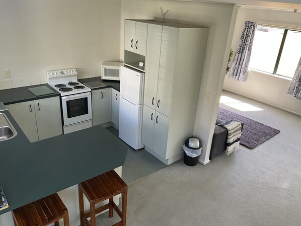 A compact kitchen with a stove, oven, microwave, and white cabinets. There is a small wooden stool and a trash can.