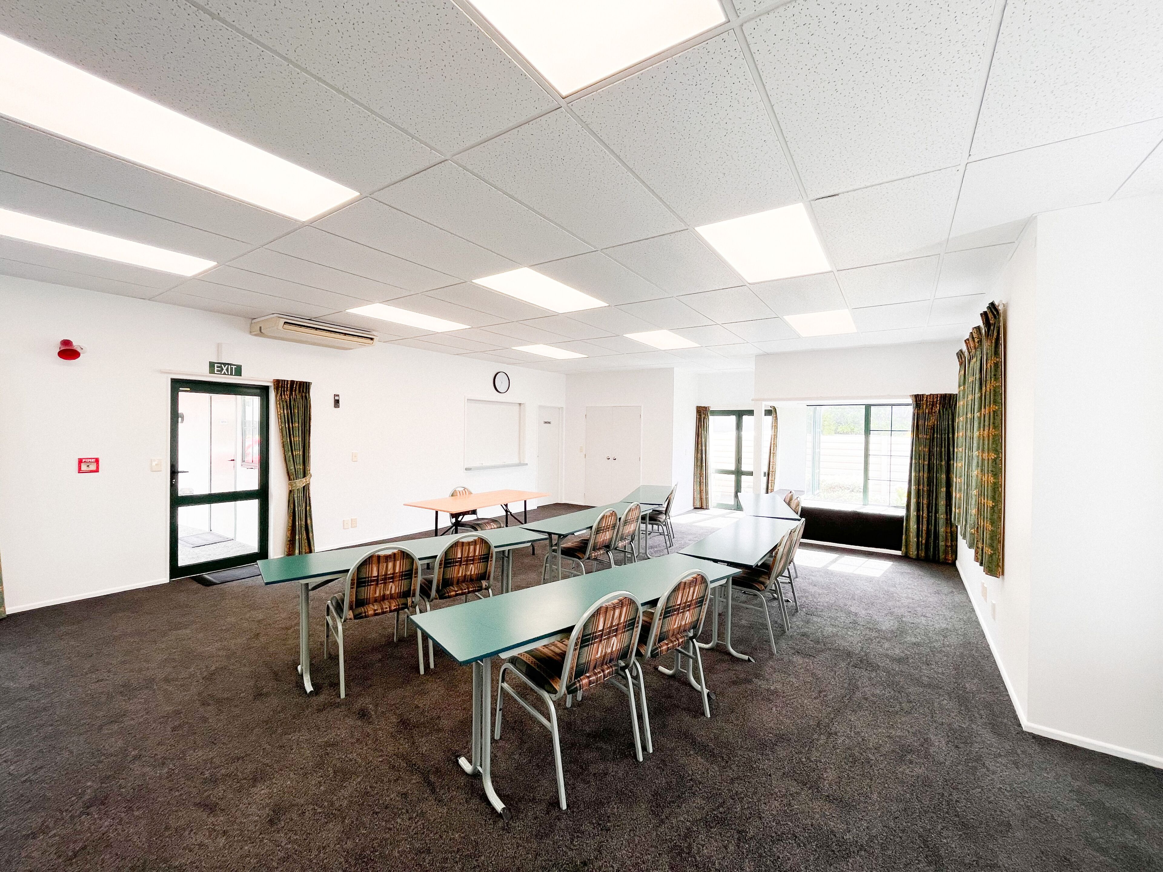 A conference room with a long table, chairs, carpeted floor, white walls, ceiling lights, exit door, and windows with curtains.