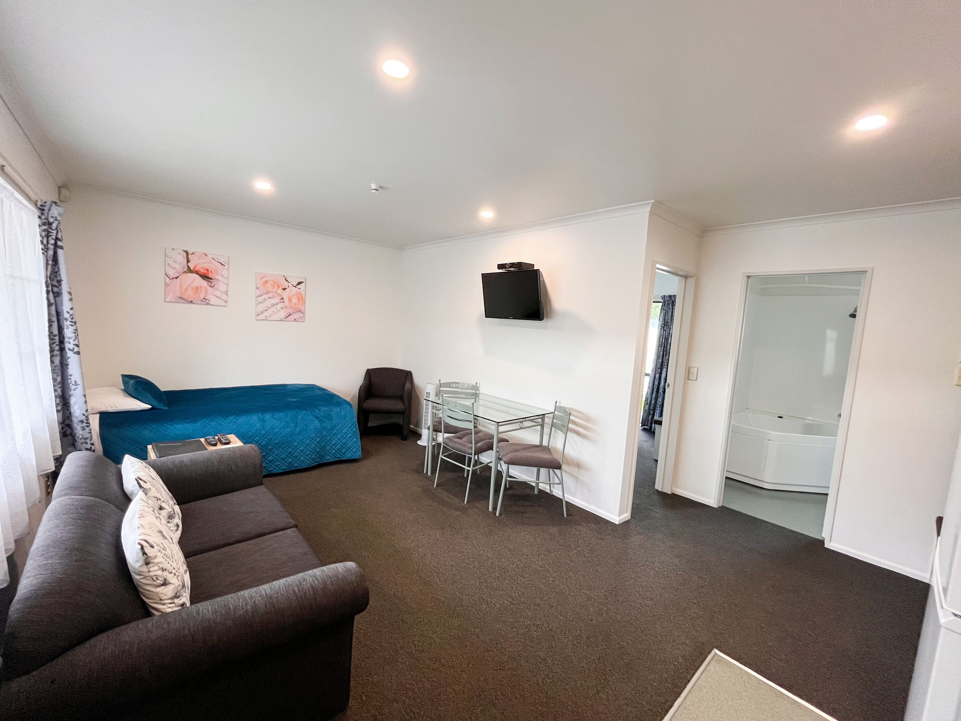 A compact room with a bed, a sofa, a small table with chairs, a TV mounted on the wall, and a door leading to a bathroom.