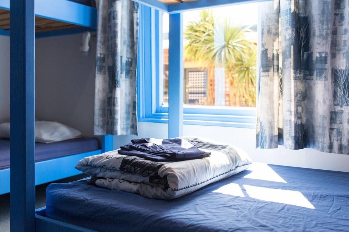 Une chambre avec un lit superposé, une fenêtre avec des rideaux et une vue sur les palmiers à l’extérieur.