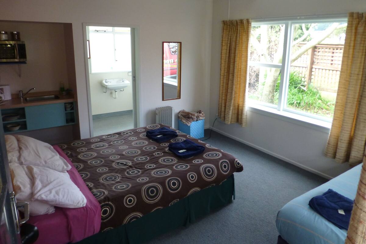 Une chambre à coucher avec un lit, un bureau, une fenêtre avec des rideaux et une salle de bain visible à travers une porte ouverte.