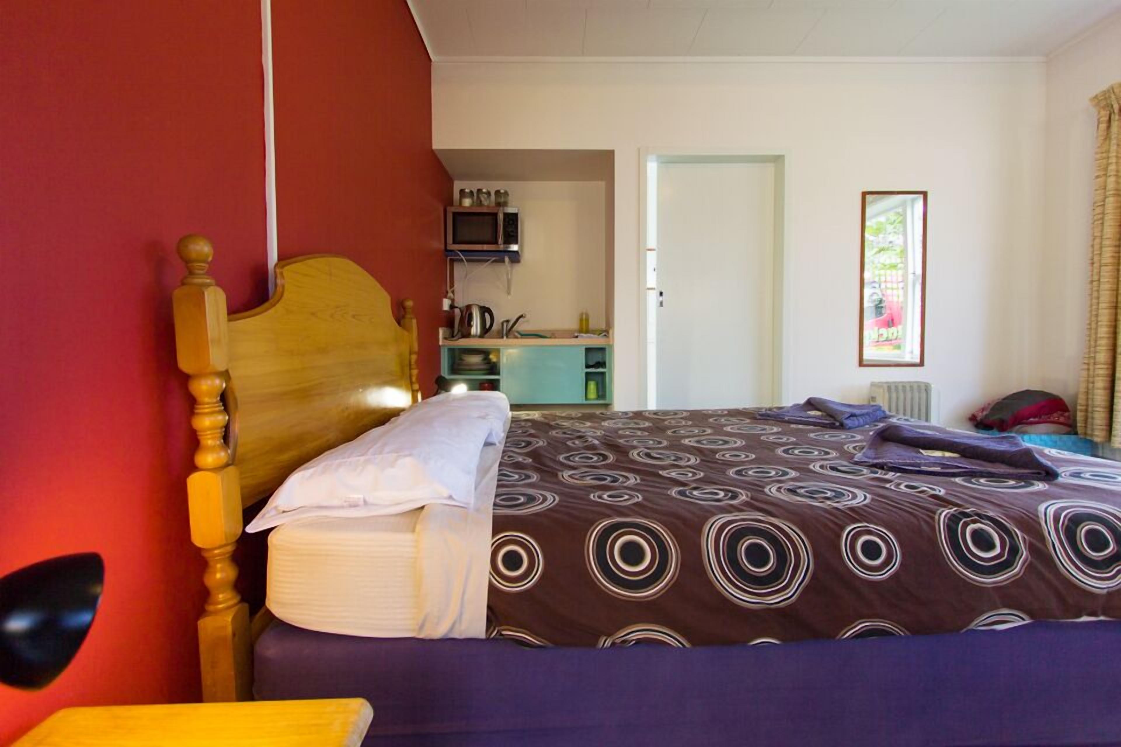 A room with a bed, a wooden headboard, a small table, a microwave, and a kitchenette in the background.
